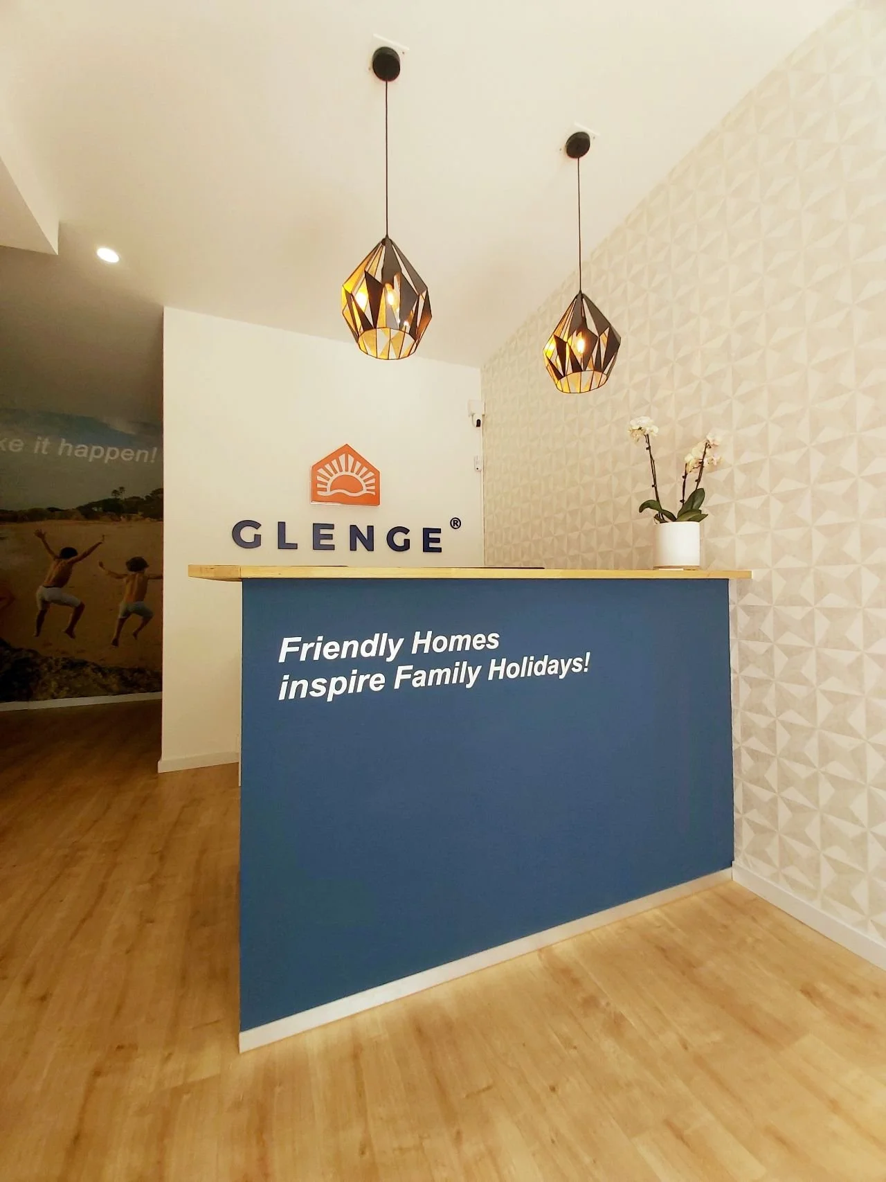 Projeto de Design de Interiores/Remodelação.
Balcão de recepção da Glenge Holiday Rentals com cor corporativa, logo em parede frontal, duas luminárias pendentes.
À esquerda, parede com impressão grande formato de uma família na praia, transmitindo o 
