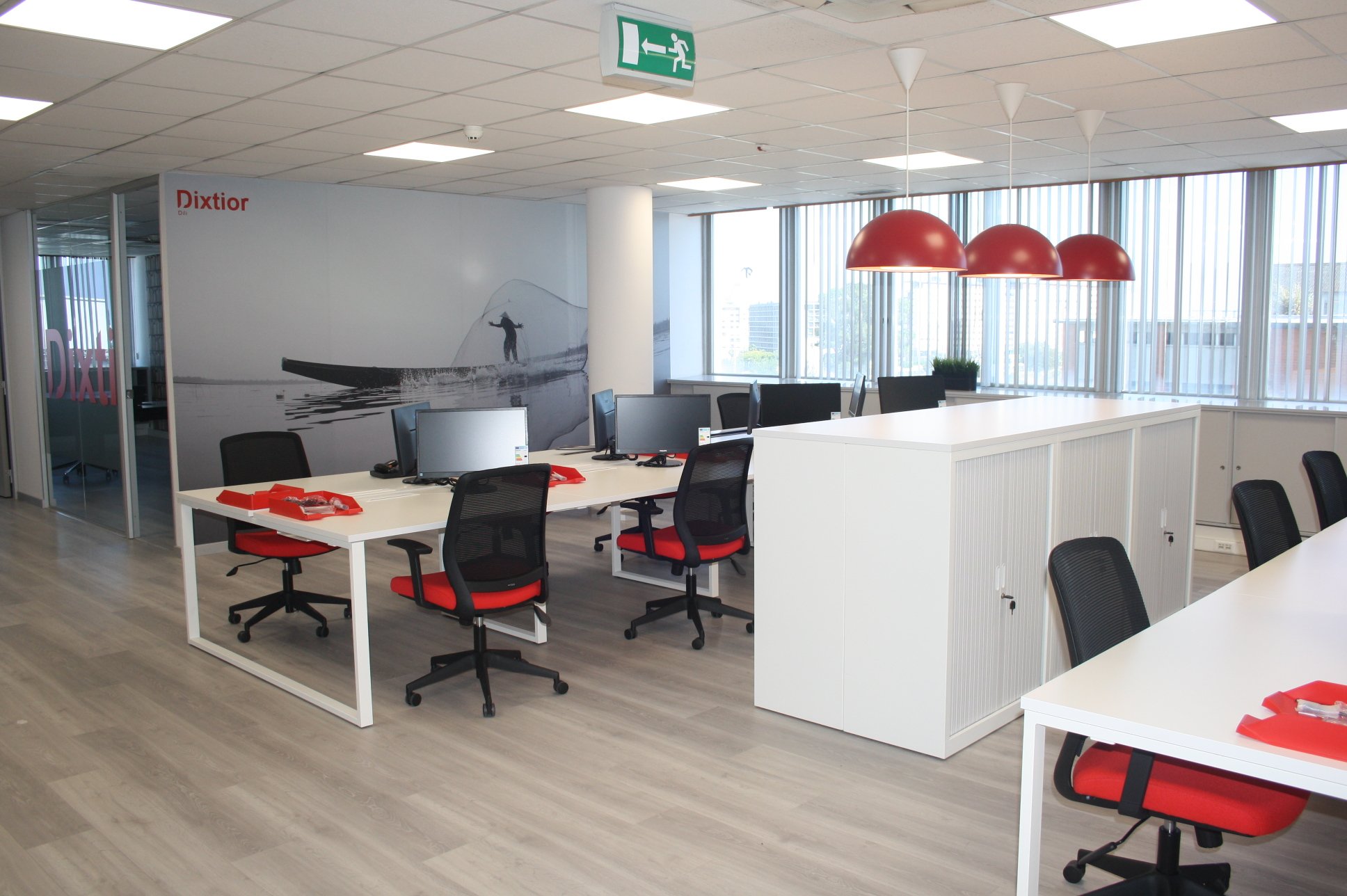 Escritório com mesas brancas, cadeiras pretas com assentos vermelhos, computadores, luminárias vermelhas e parede com mural de uma pessoa em um barco de pesca de alga