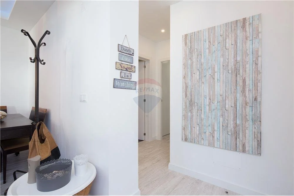 Decoração de parede com painéis de madeira clara, uma placa de madeira com palavras em inglês, uma espreguiçadeira com itens de decoração, um cabideiro de ferro preto com roupas e uma mesa redonda com objetos decorativos em um ambiente de interior.