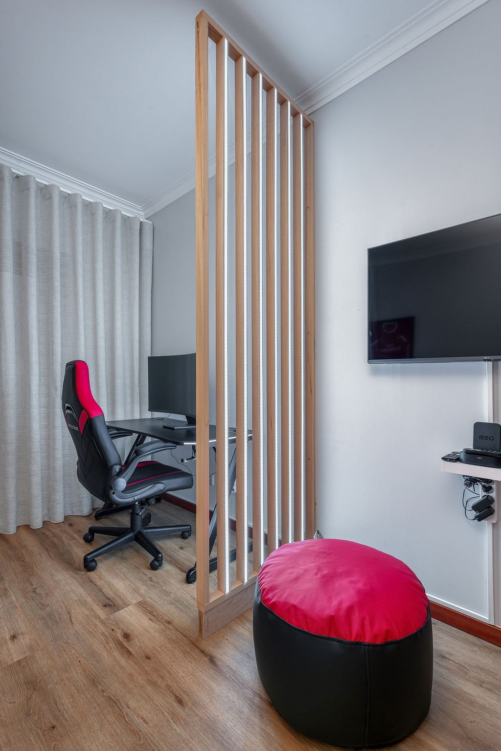 Sala de estar com parede branca, televisão de tela plana na parede, poltrona de jogos preta e vermelha, estante com controle remoto e roteador, cortina bege ao fundo, divisória de madeira entre a área de trabalho e a área de descanso, e pufe preto e vermelho no chão de piso de madeira.