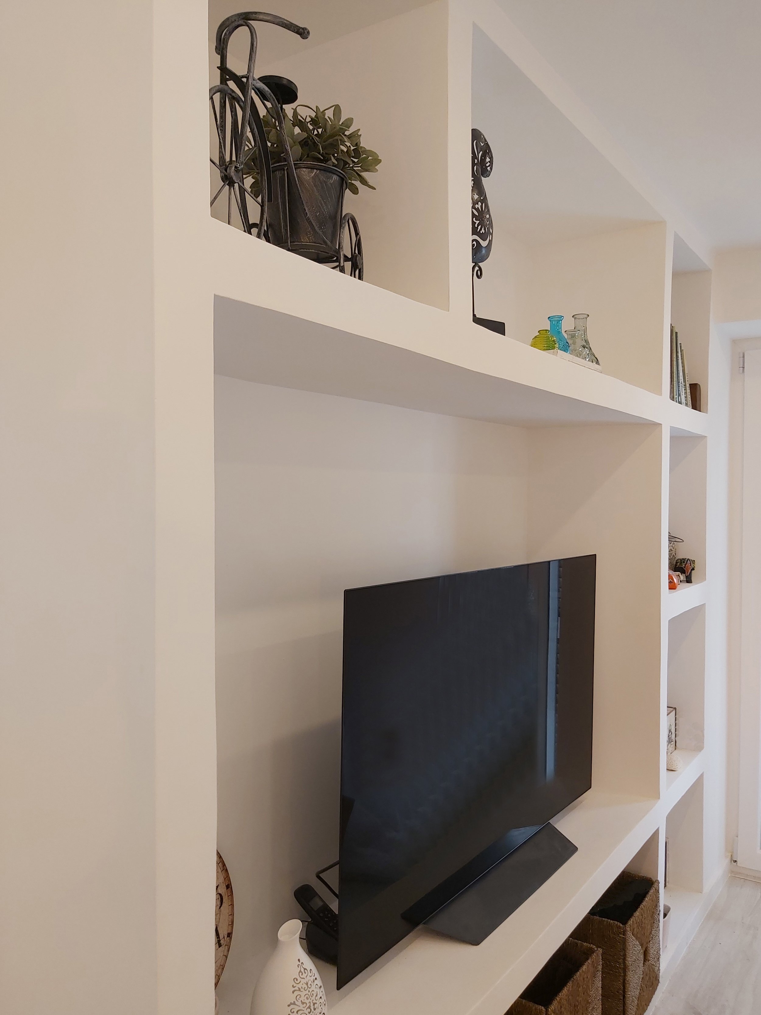 Tela de televisão em uma parede branca com estantes de nichos acima, decoradas com livros, vasos de vidro, uma bicicleta decorativa e uma planta em um vaso metálico.