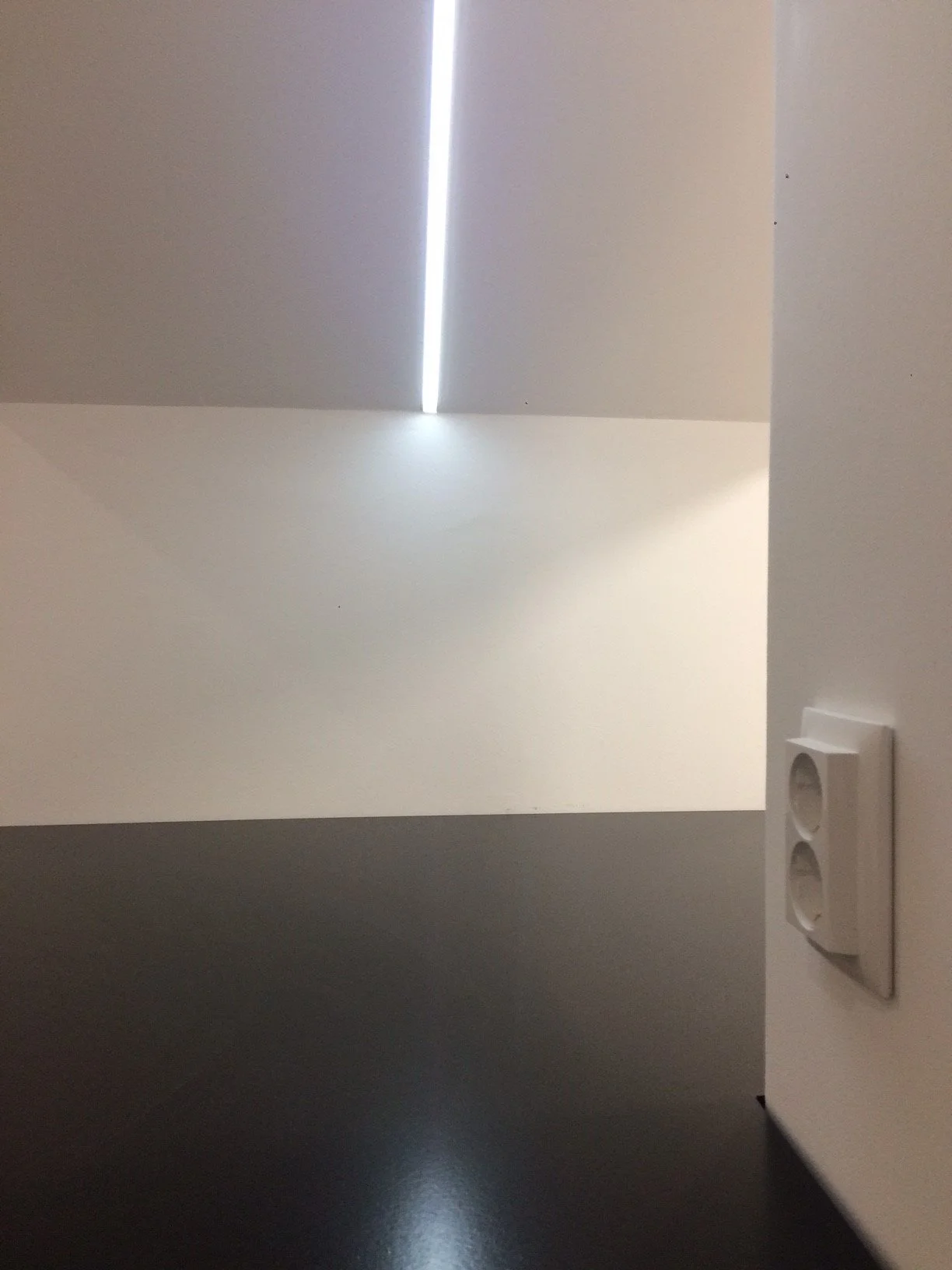 Interior de uma sala com iluminação natural, parede branca, teto alto com uma faixa de luz e uma tomada de energia na parede à direita, piso preto brilhante.