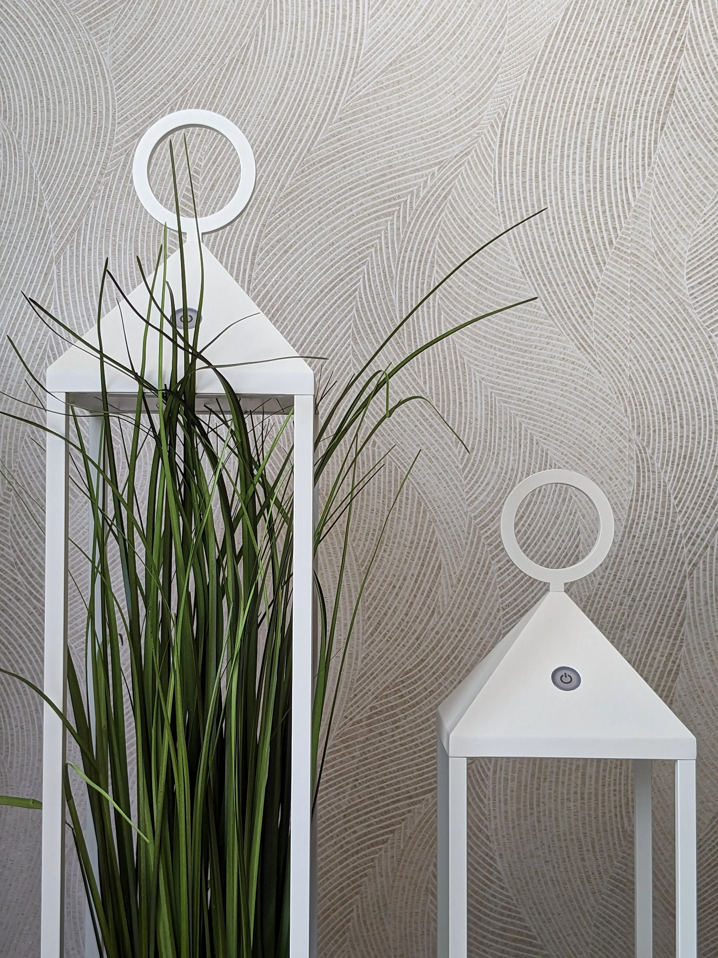 Decoração com duas luminárias brancas modernas e uma planta com folhas longas e finas, em frente a uma parede com papel de parede colorido de padrão de folhas pequenas.