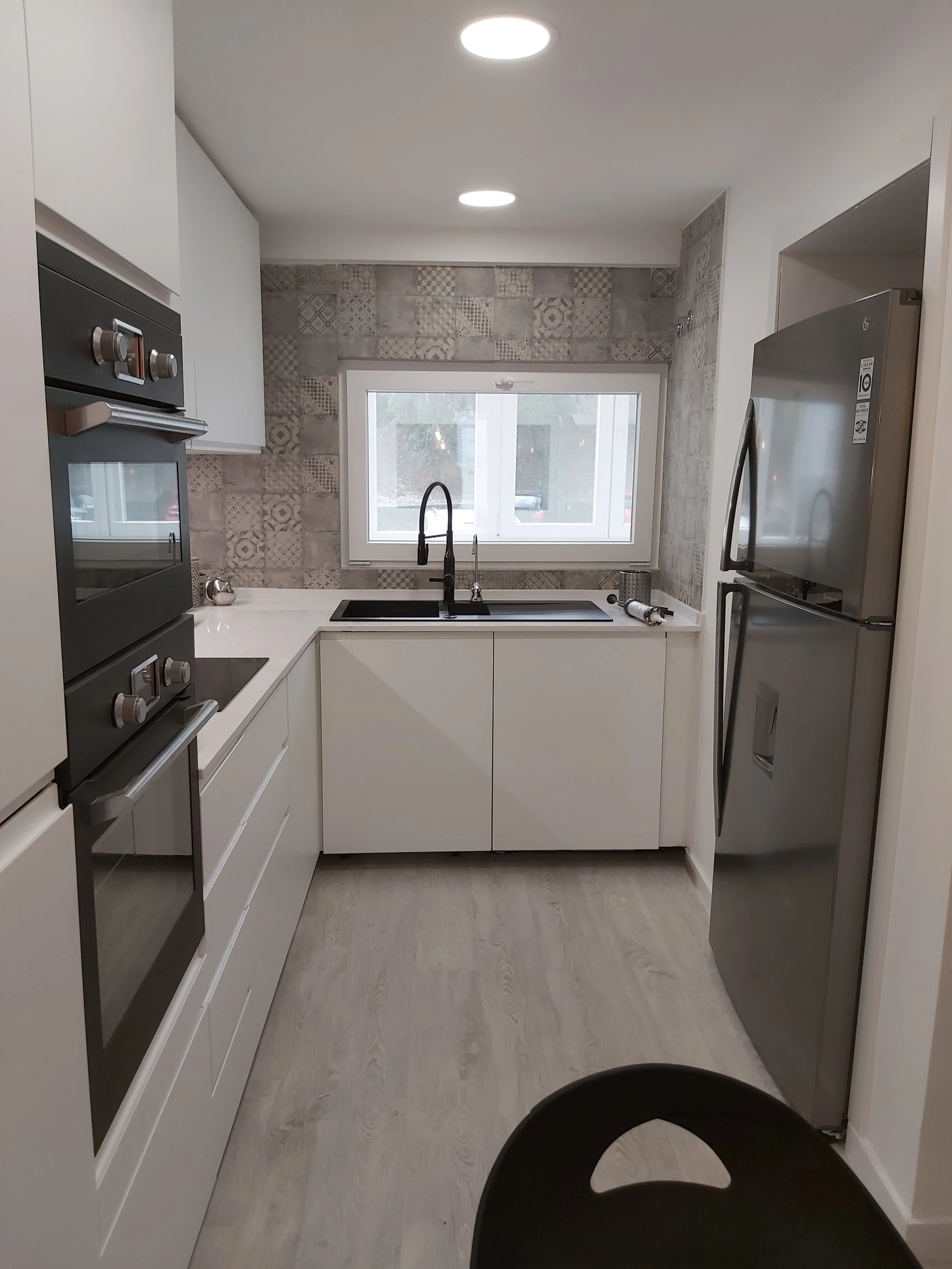 Cozinha moderna com armários brancos, forno embutido, pia com torneira preta, janela ao fundo, parede revestida com azulejos decorativos em tons neutros, geladeira de inox à direita e um balcão com utensílios à direita.