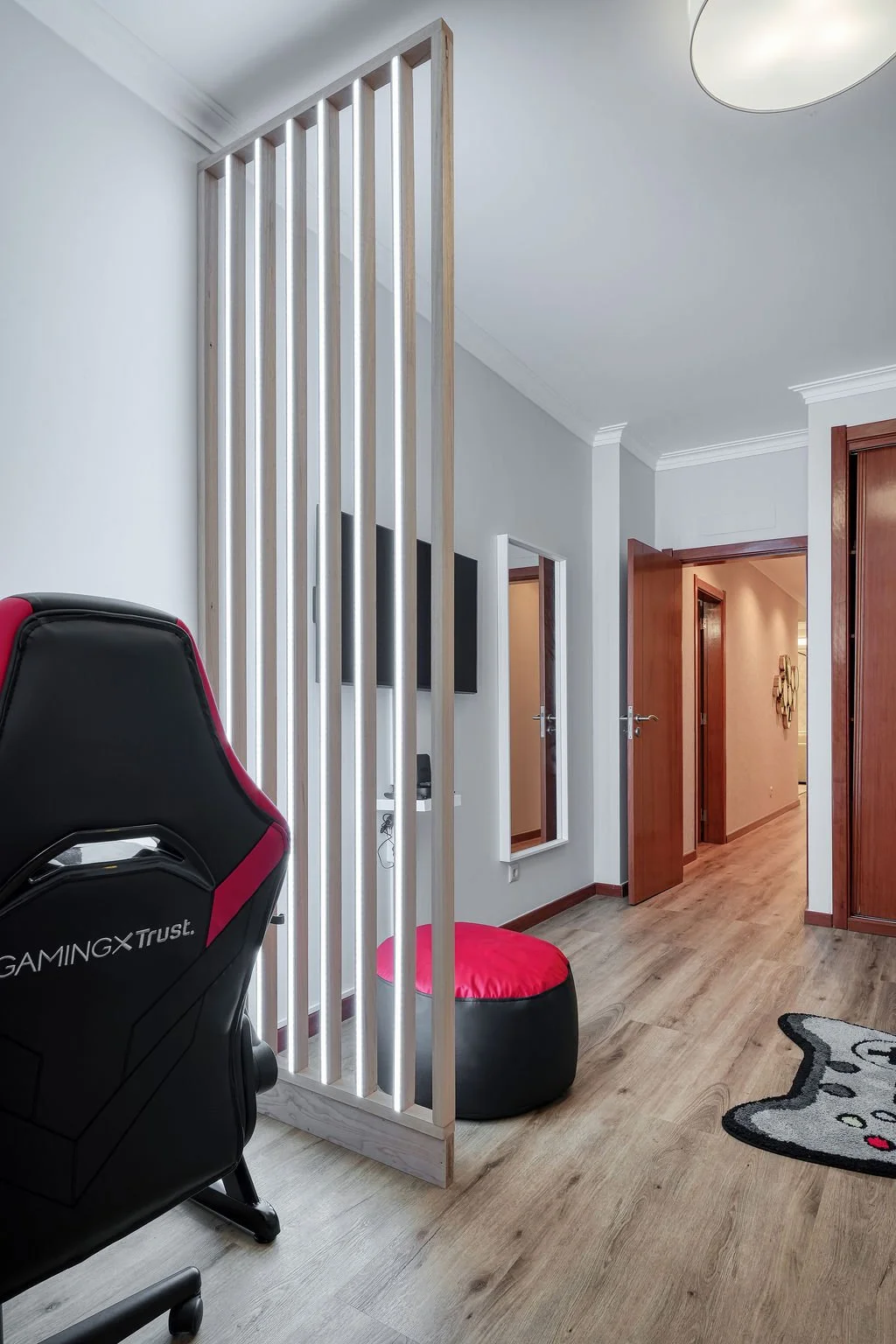 Cadeira de gaming preta com detalhes vermelhos, uma almofada vermelha, parede clara com um espelho de corpo inteiro, uma porta aberta conduzindo a outro cômodo, piso de madeira, parte de tapete com desenho de gato