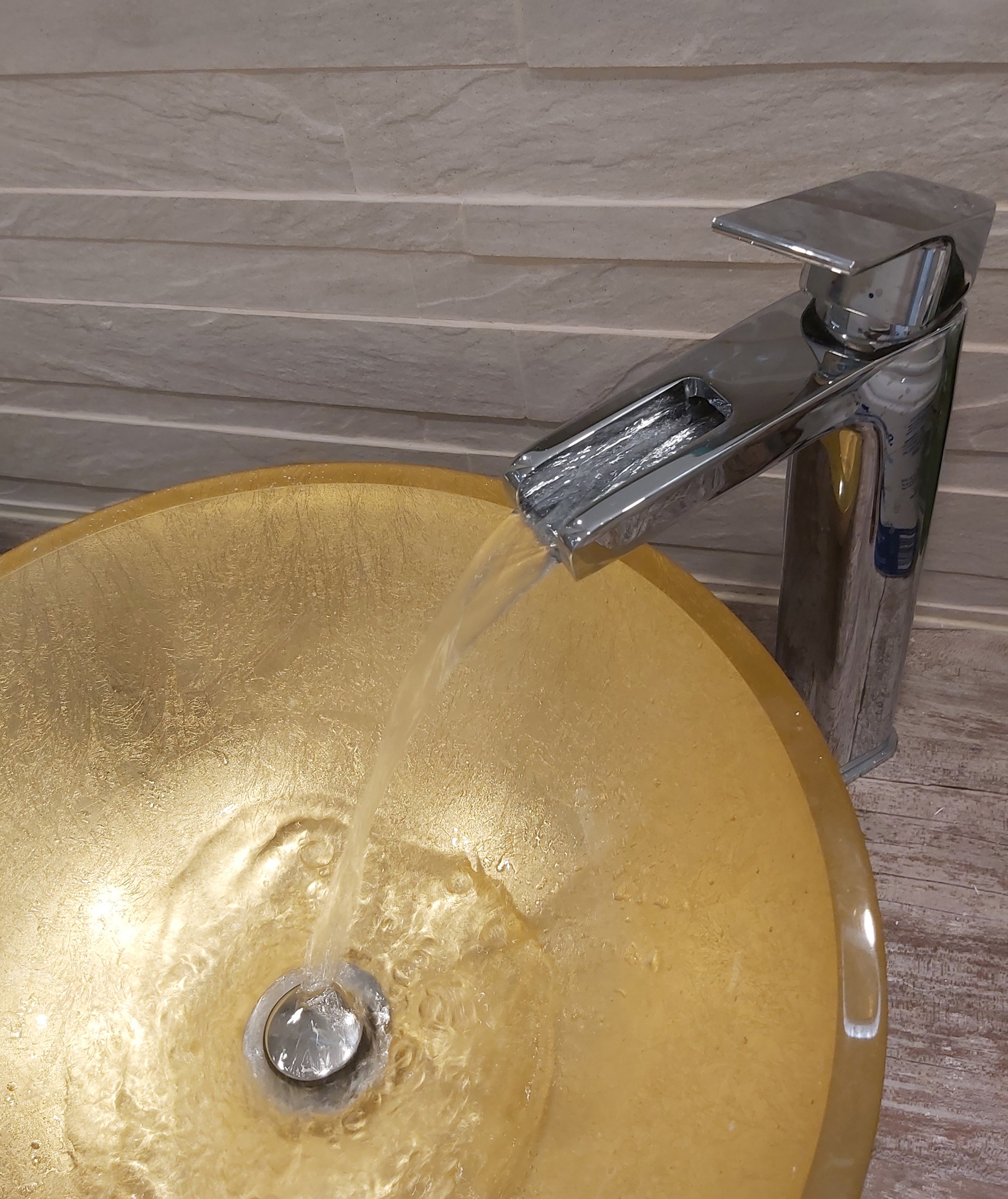 Torneira de metal dourada, com água jorrando, instalada acima de uma pia dourada com textura brilhante, paredes de tijolos em tom claro ao fundo.