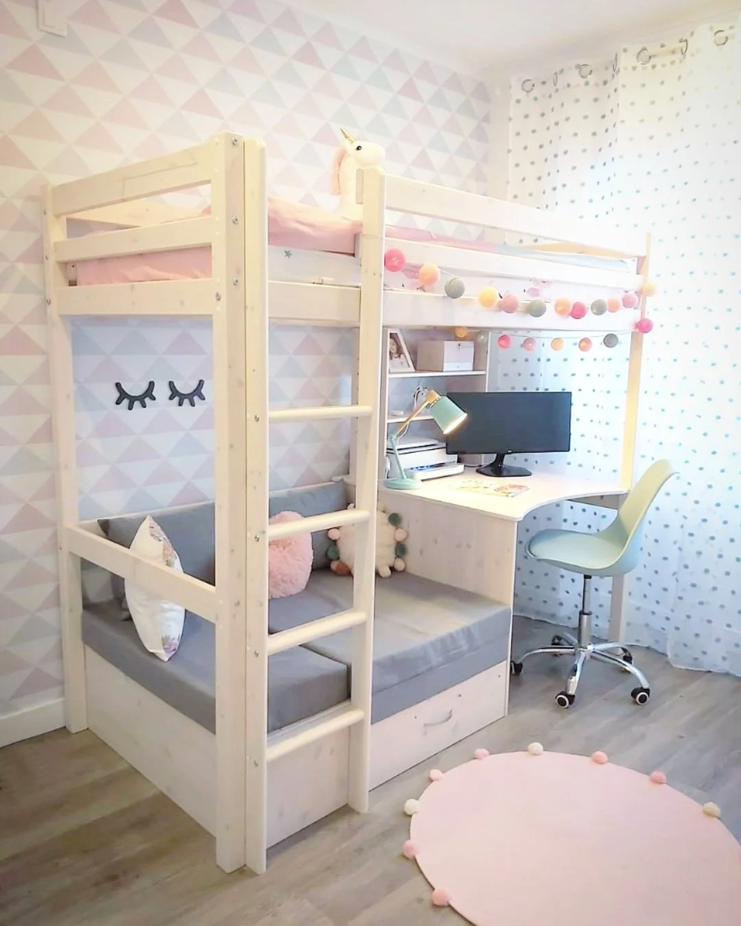 Quarto infantil decorado com cama de beliche de madeira clara, almofadas fofas, cortinas com estampa de bolinhas, parede com adesivo de olhos fechados, tapete rosa com pompons, área de estudo com computador e luminária, cadeiramede de escritório, pai