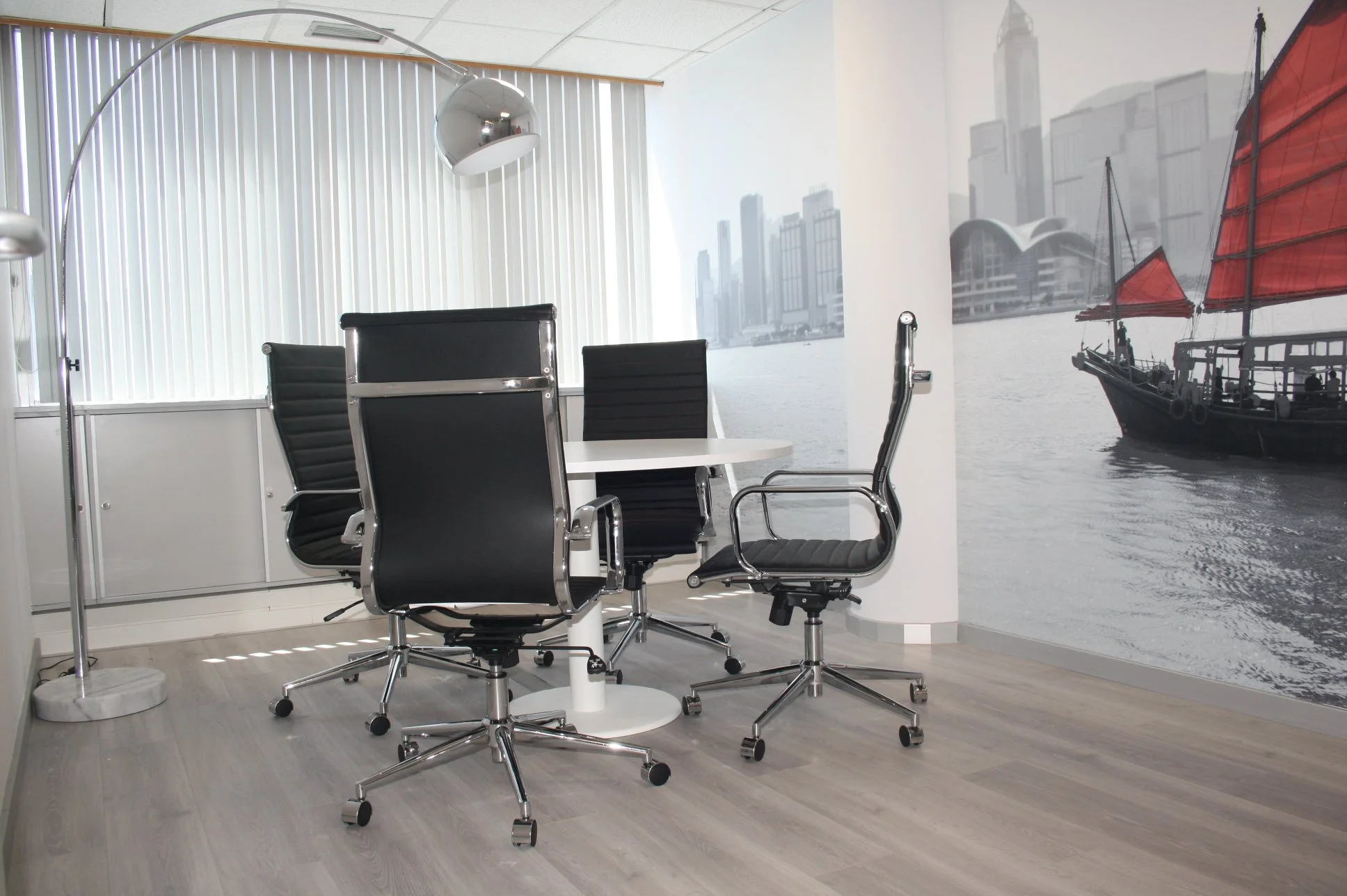 Sala de reunião com mesa branca, cadeiras pretas com estrutura de metal, maior uma frente de uma janela com persianas brancas, e uma parede com mural de cidade com prédio e barco a vela vermelho.