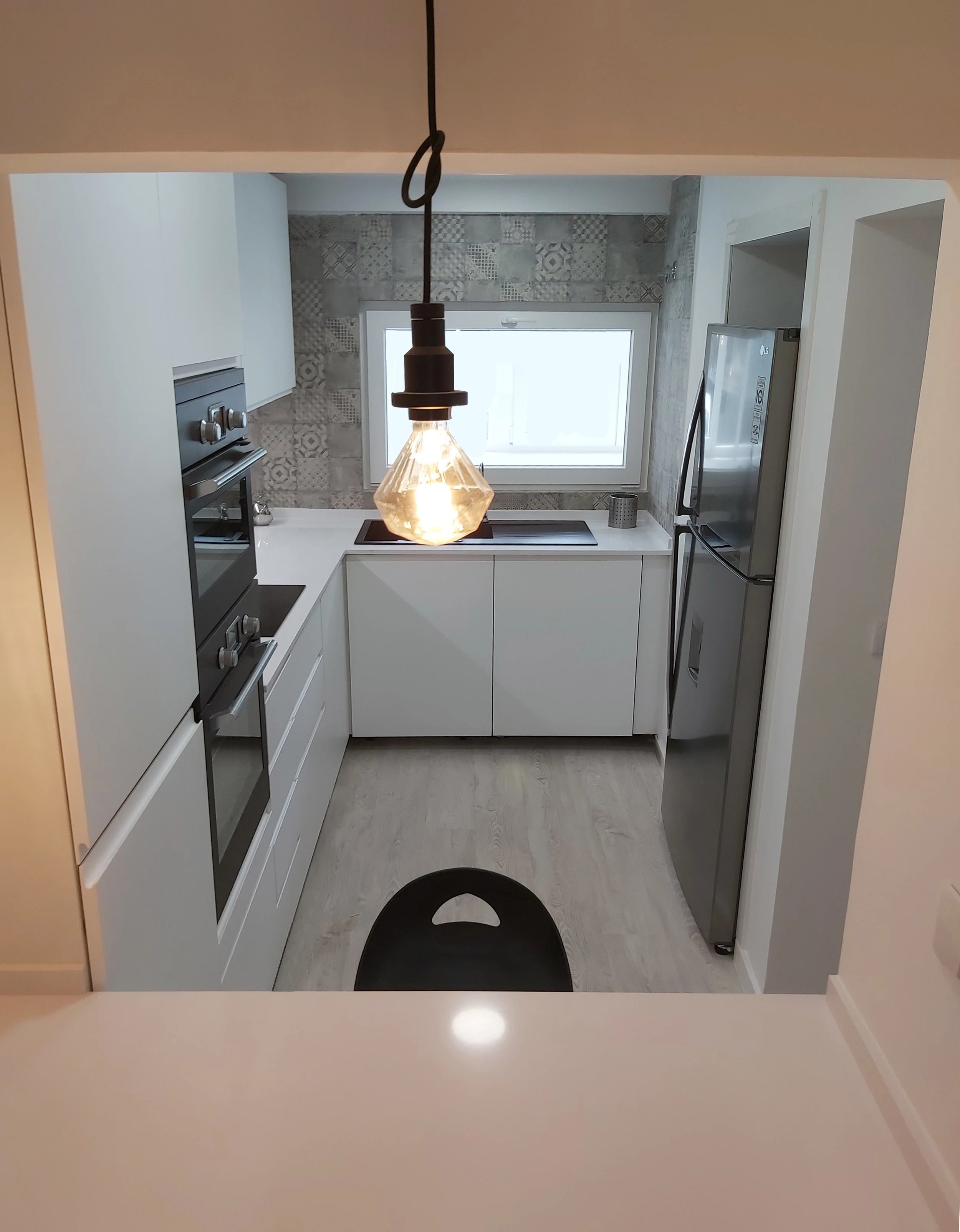 Cozinha moderna com armários brancos, geladeira de aço inox, forno, cooktop, janela, iluminação suspensa com lâmpada de filamento exposta e piso de madeira clara.