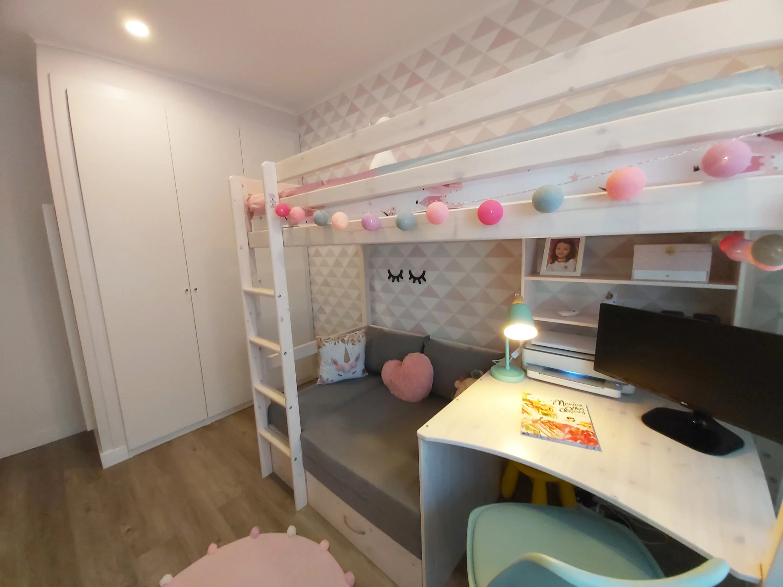 Quarto infantil decorado com cama suspensa, almofadas de unicórnio e coração, parede com papel de parede de triângulos, escrivaninha com computador, prateleiras com fotos e objetos, luz de leitura, cadeiras coloridas e lindas luzes decorativas