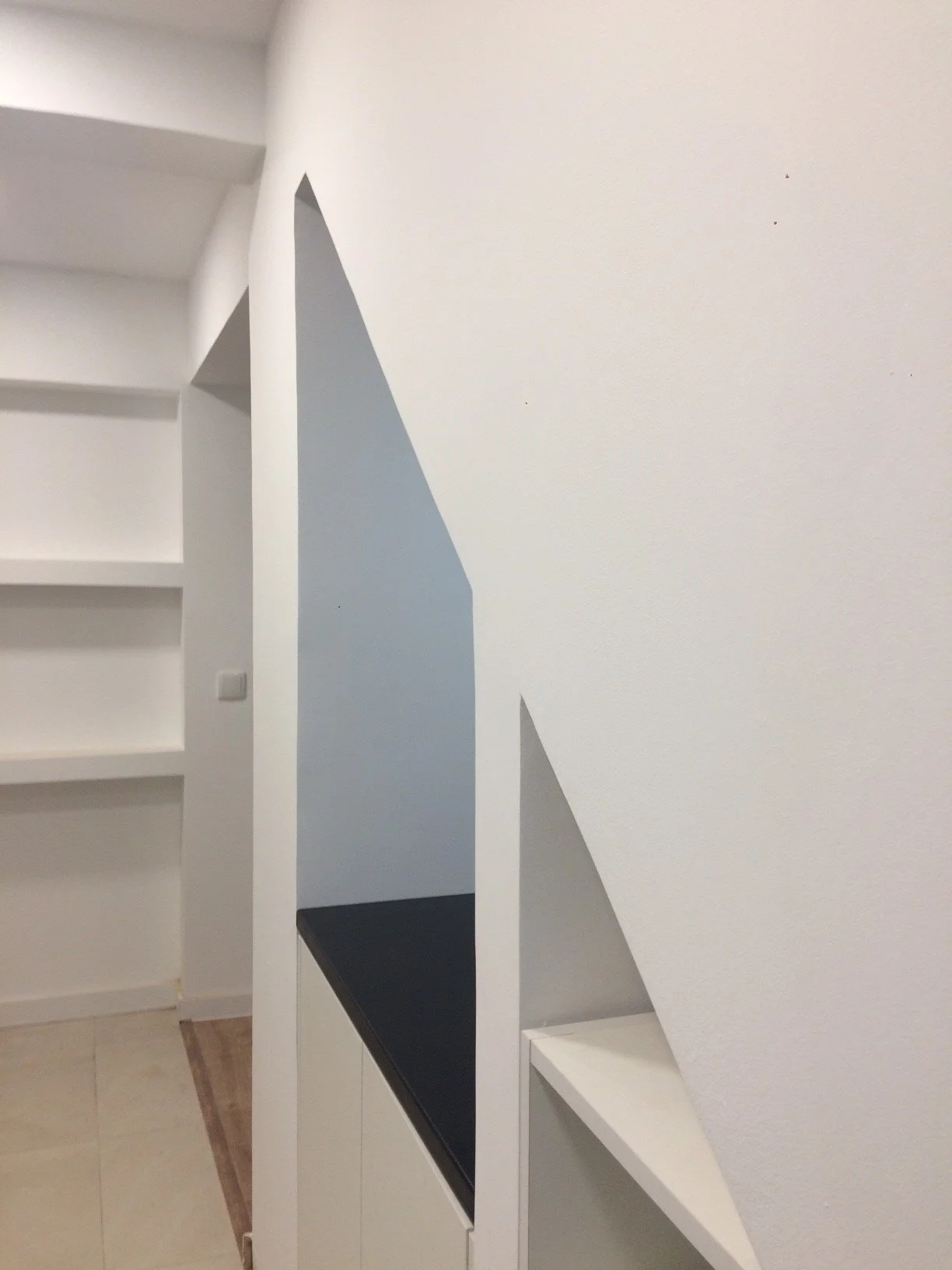 Parede branca com detalhes em azul escuro e uma porta de correr preta, parte de uma sala ou corredor com prateleiras brancas.