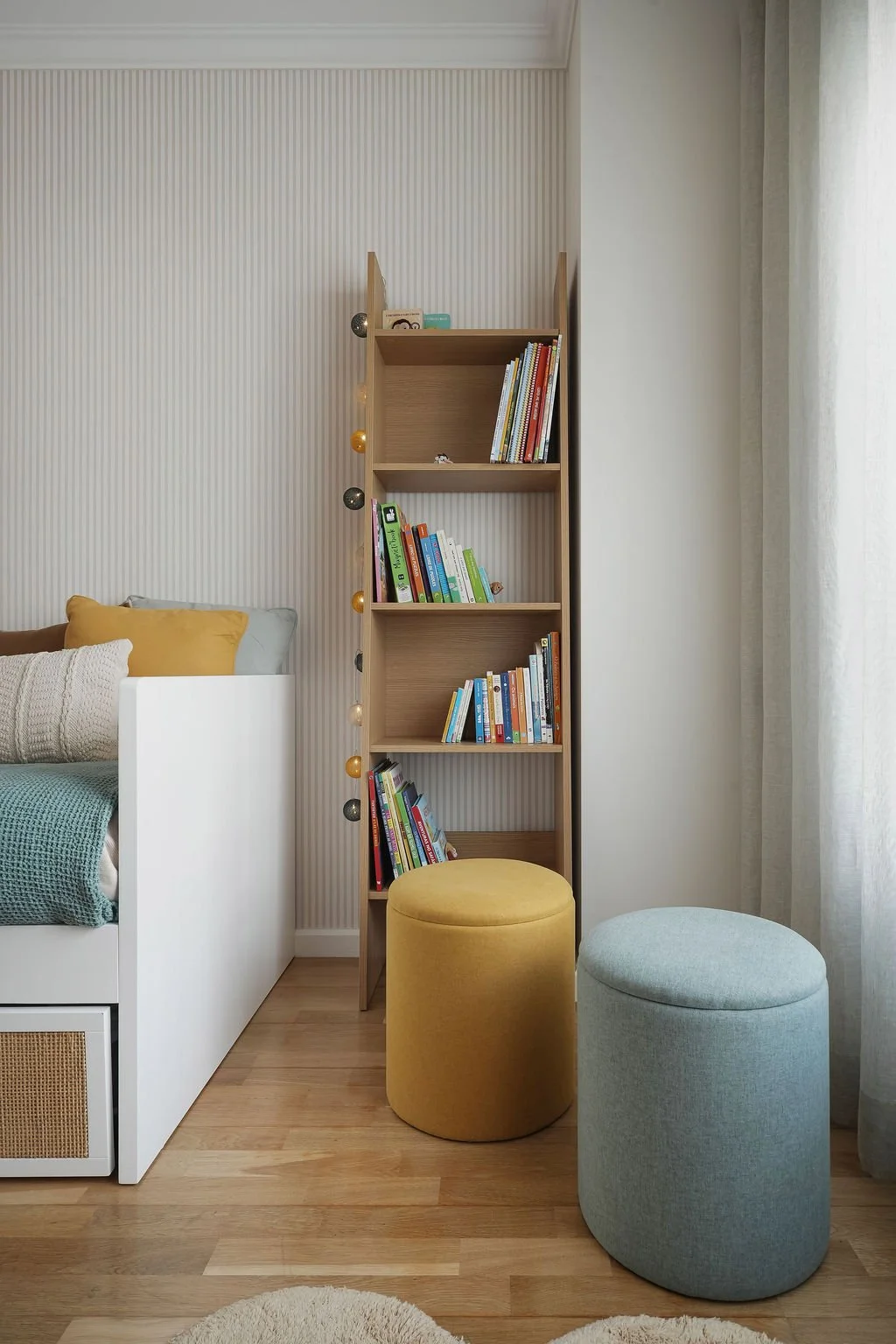 Estante de madeira com livros e luzes decorativas ao lado de uma cama com travesseiros amarelo, cinza e por cima de um tapete redondo na sala