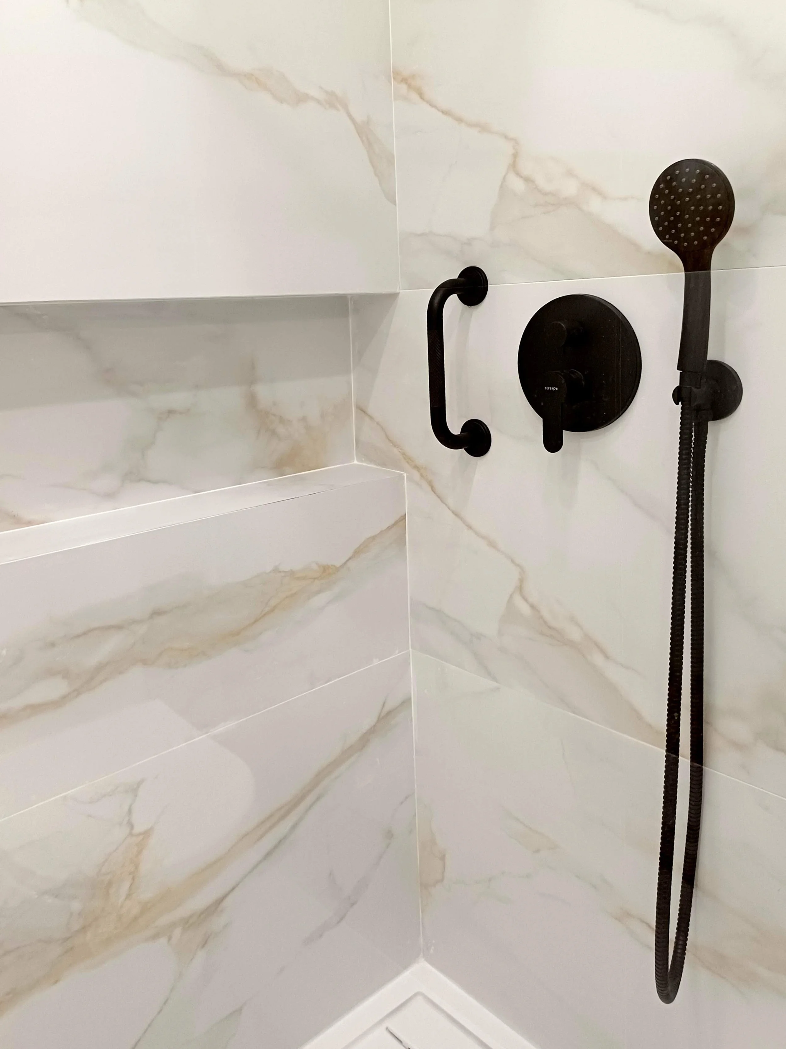 Chuveiro preto com barra de suporte e controle de temperatura em banheiro com revestimento de mármore branco com veias douradas.