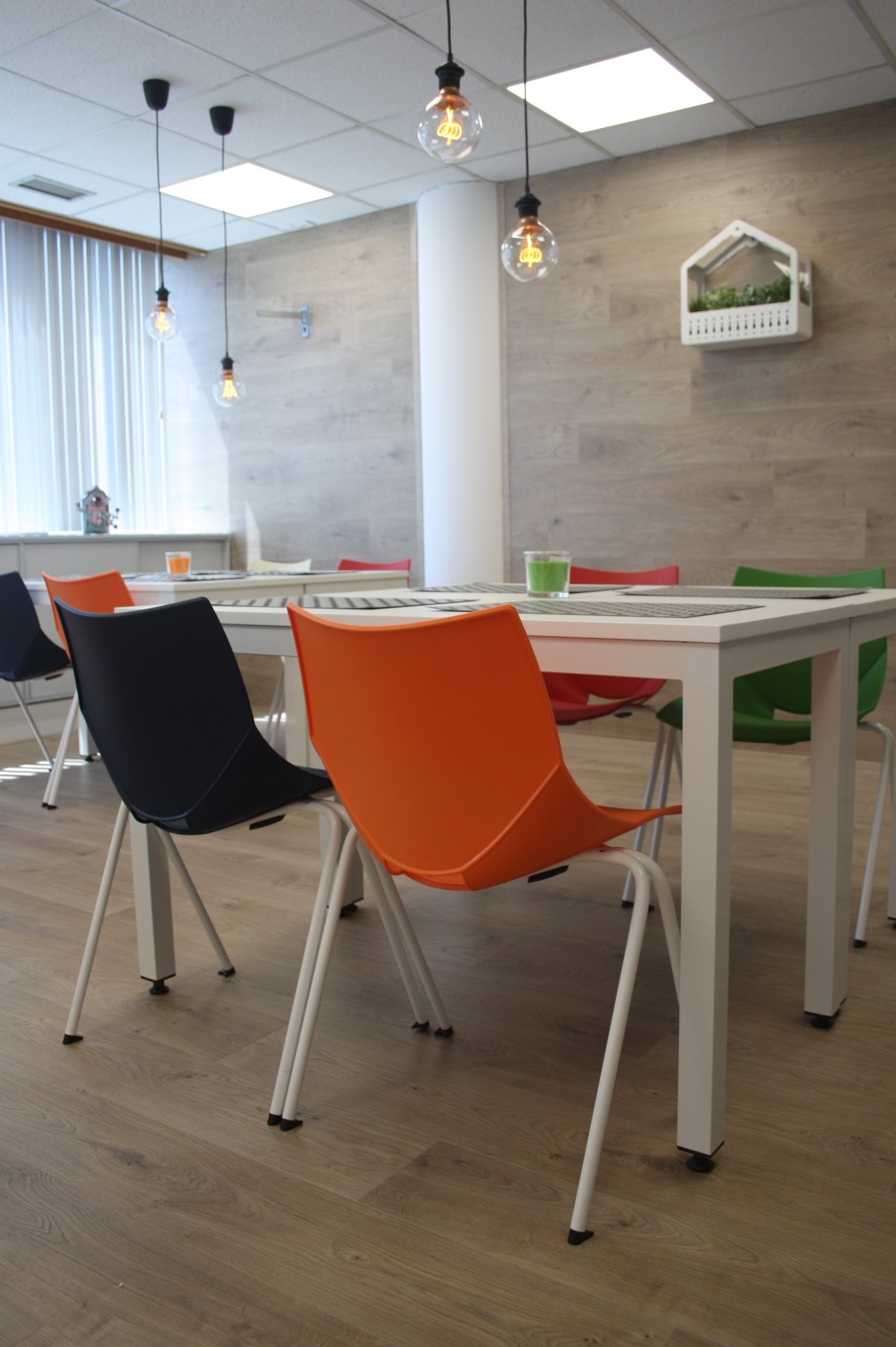 Sala de reunião com mesa branca, cadeiras de plástico em cores preto, laranja, vermelho e verde. Iluminação com luminárias de lâmpadas aparentes. Janela com cortina de persianas, parede de madeira clara com uma prateleira branca decorativa com planta