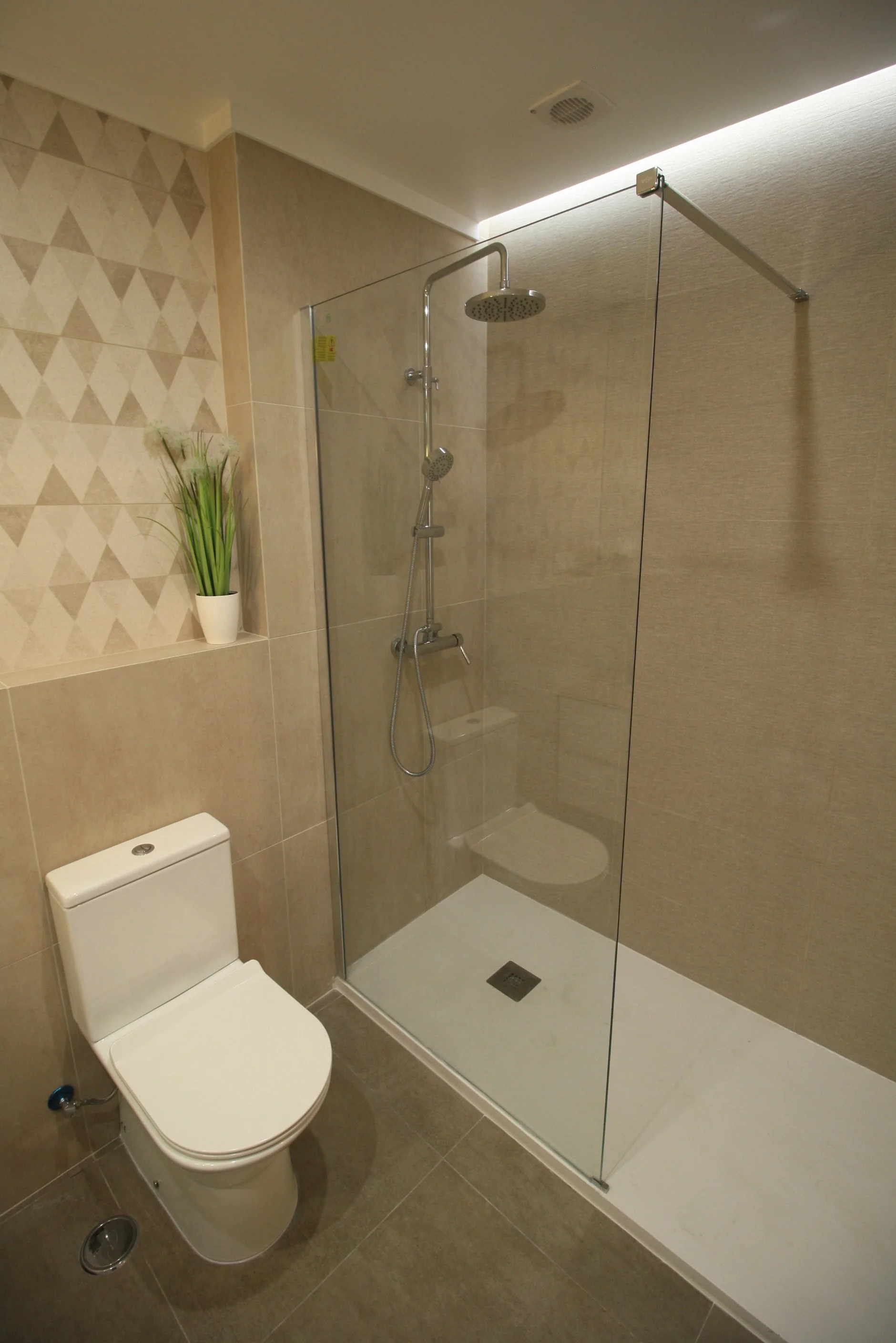 Banheiro moderno com vaso sanitário, ducha com vidro e revestimentos em tons de bege, decorado com planta em vaso branco.