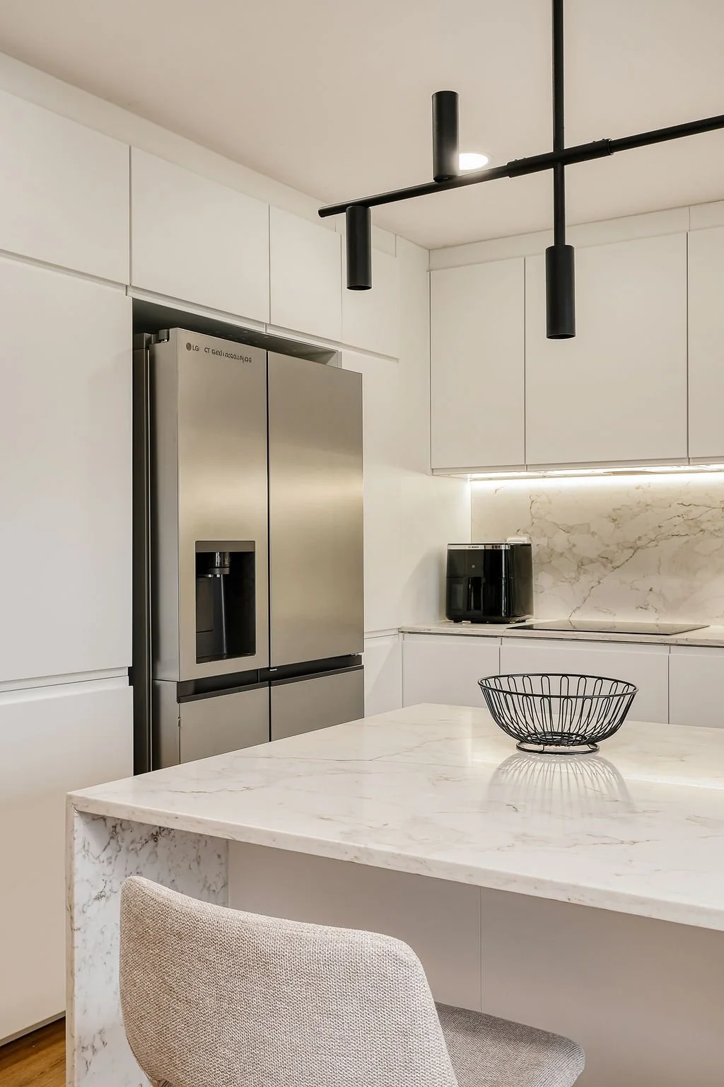 Cozinha moderna com armários brancos, geladeira de aço inox, bancada de mármore, cafeteira preta e uma mesa de mármore com uma cadeira beijada