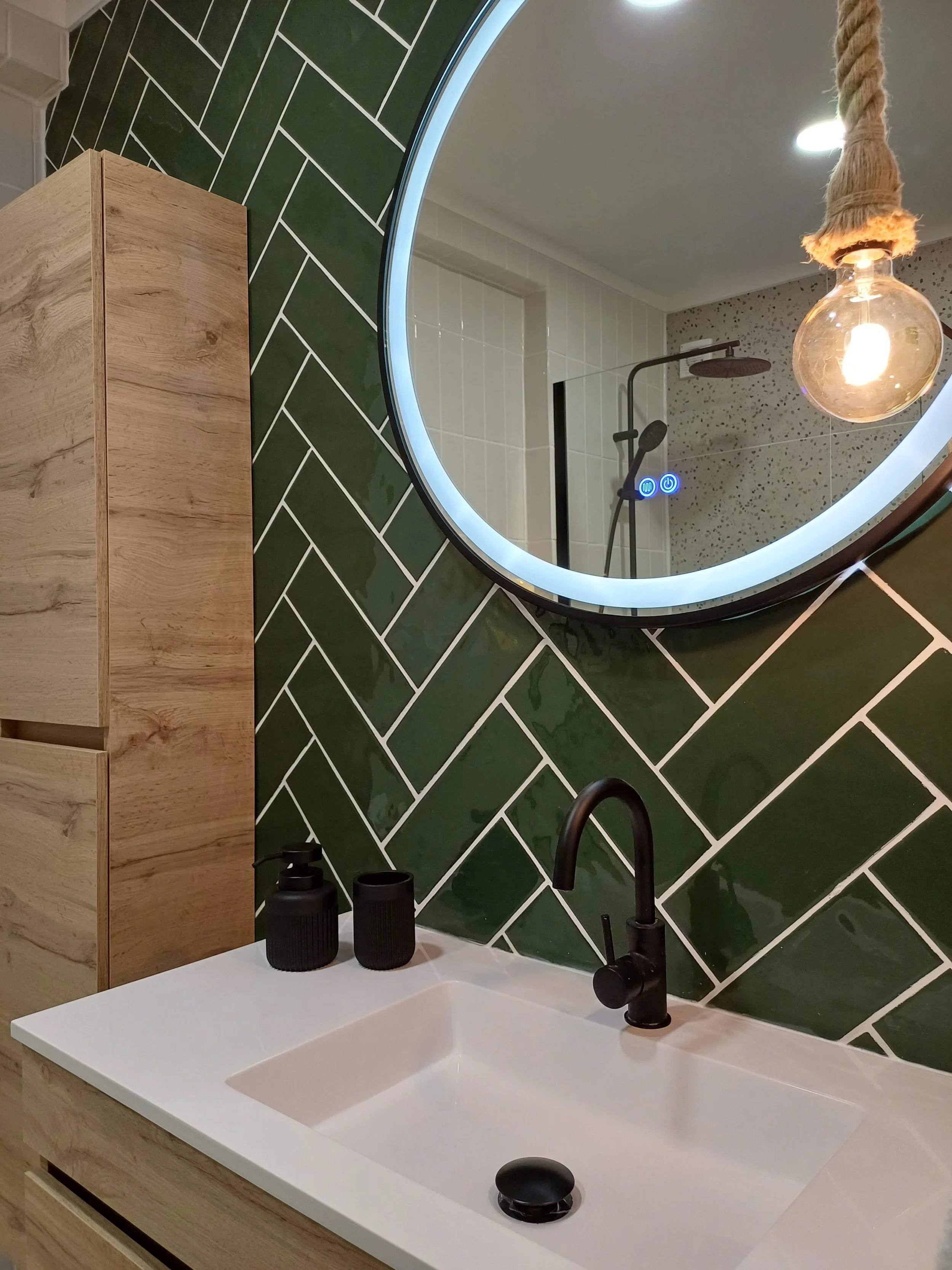 Espelho redondo com iluminação ao redor refletindo um chuveiro no banheiro. Pia branca com duas dispenser de sabonete preto e um vaso de escova de dente preto. Parede revestida de azulejos verdes em padrão diagonal. Armário de madeira na esquerda. Fi