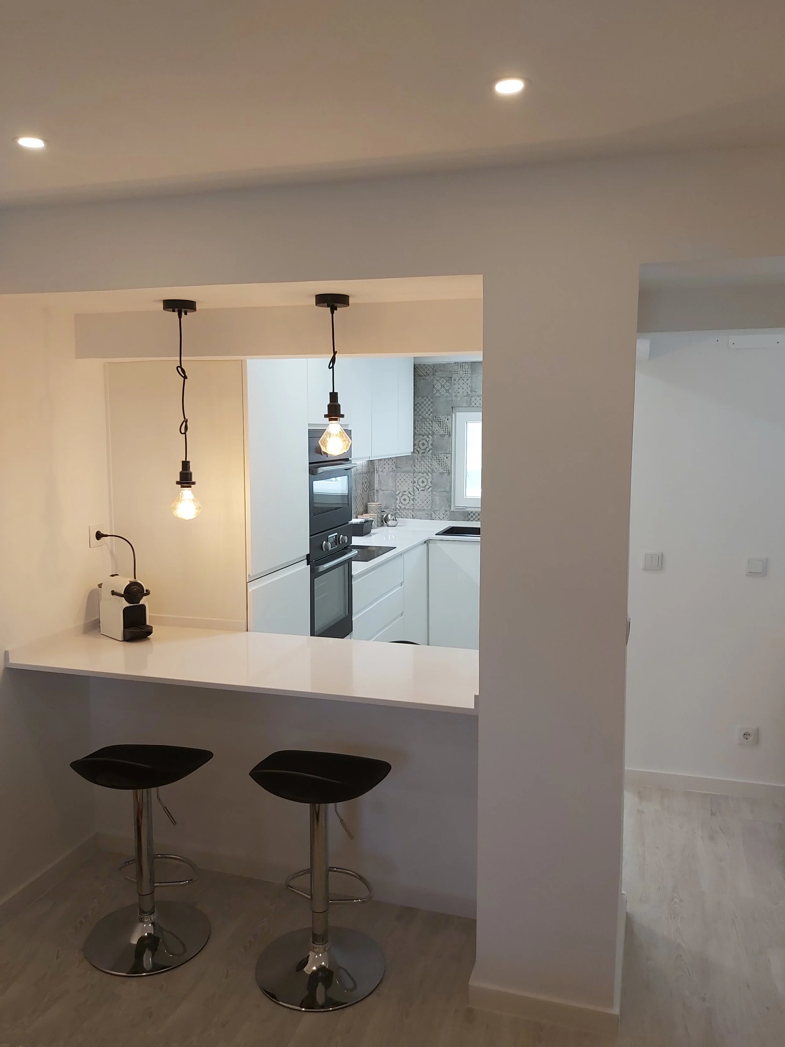 Cozinha moderna com iluminação pendente, bancada branca, dois bancos altos com assento preto, eletrodomésticos embutidos, parede com azulejos decorativos cinza, janela ao fundo, piso de madeira clara.