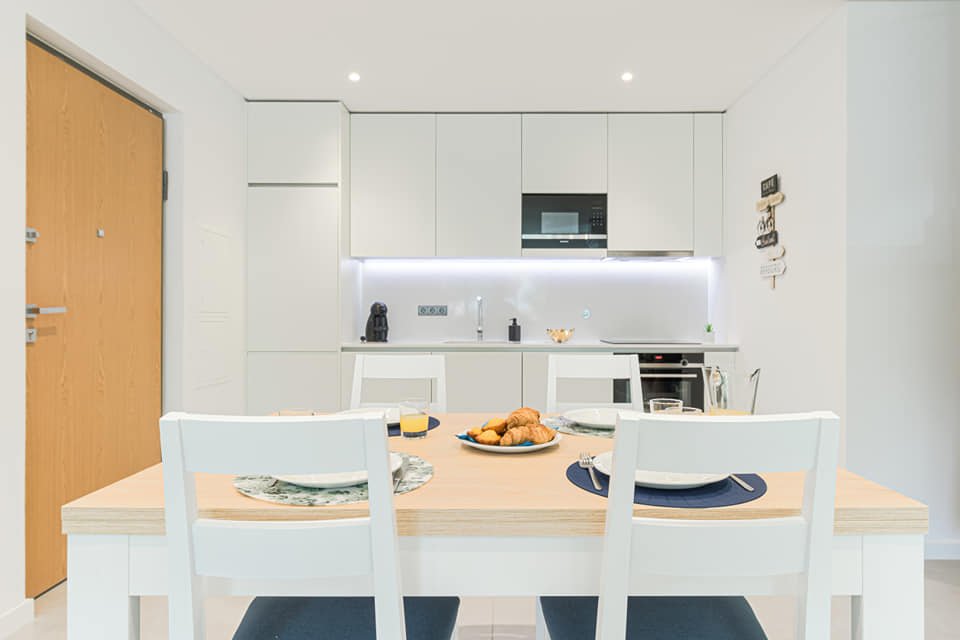 Cozinha moderna com mesa de jantar de madeira clara, quatro cadeiras brancas, e alimentos no centro. Móveis brancos e eletrodomésticos embutidos na parede branca, iluminação LED sob armários, porta de entrada de madeira ao lado esquerdo.