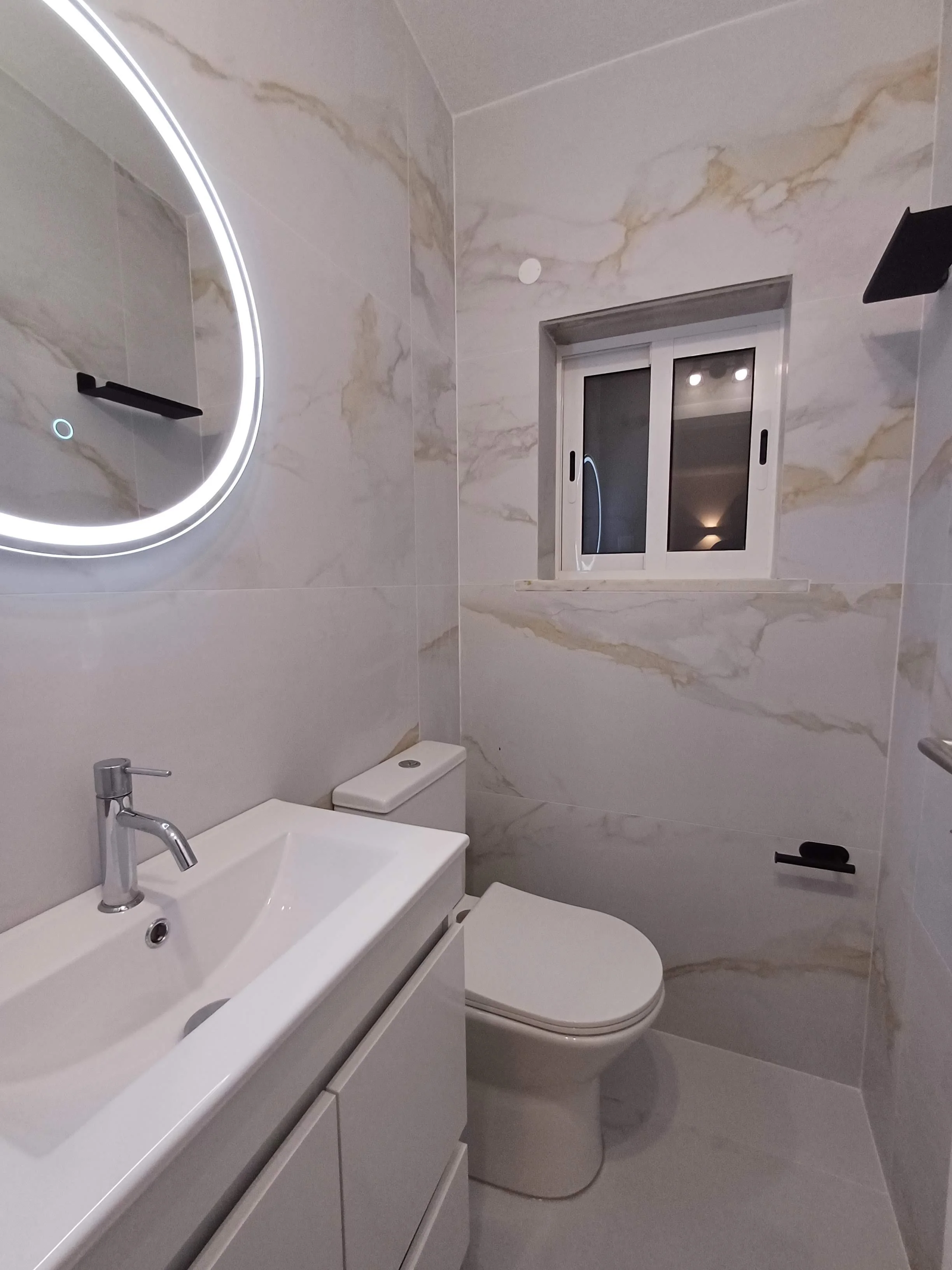 Banheiro com paredes de mármore claro, espelho com iluminação de led, vaso sanitário e pia branca, janela pequena e preta