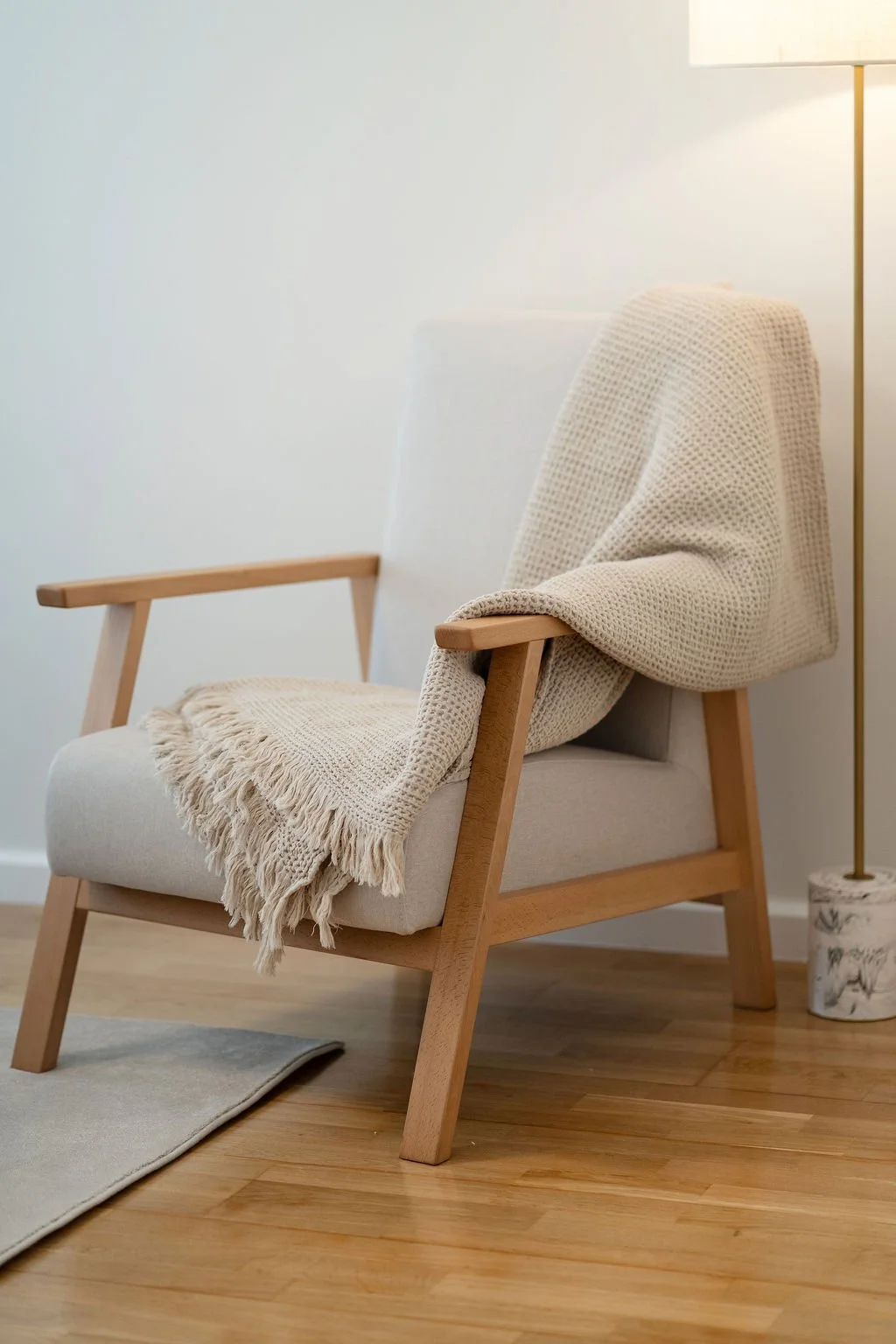 Poltrona de madeira estofada a branco, coberta por uma manta bege. Tapete cinza claro e candeeiro de pé com haste dourada e base marmoreada.