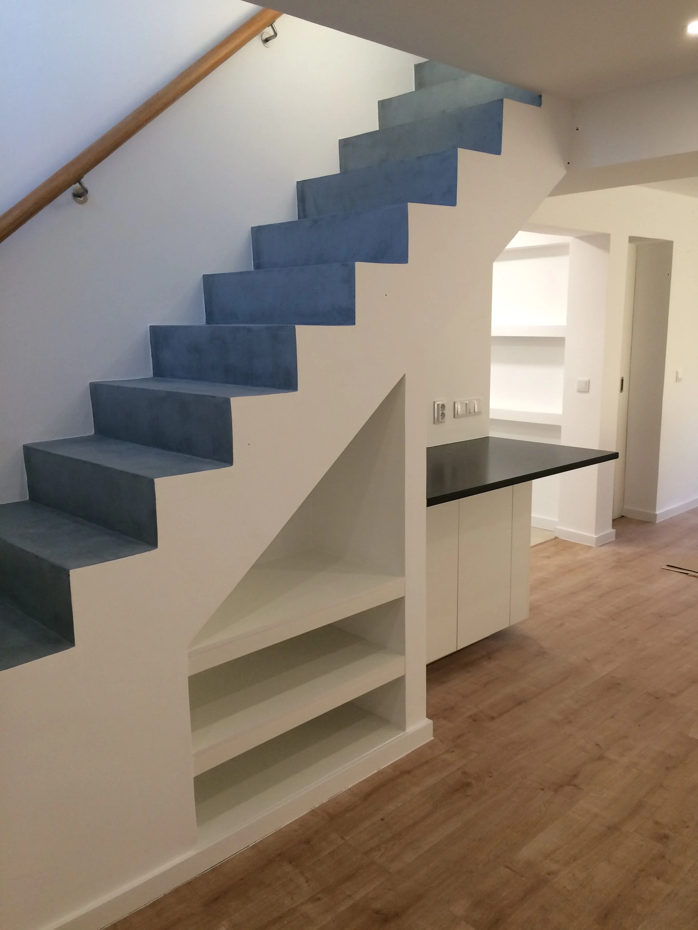 Escada de interior com degraus azuis e guarda-corpo de madeira, parede branca, prateleiras embutidas na parede, bancada preta e piso de madeira.