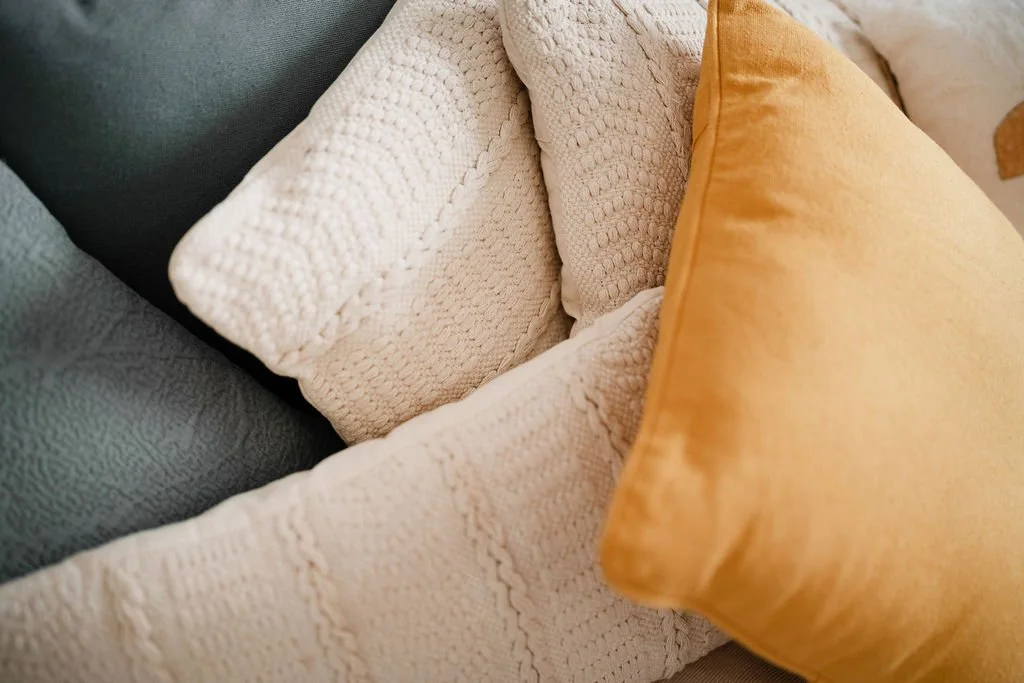 Imagem de três almofadas coloridas empilhadas, uma cinza, uma creme com textura de tricô e uma amarelo moutarde, sobre um sofá.