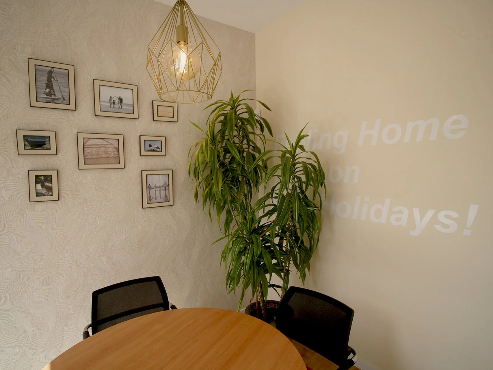 Sala de reunião com mesa de madeira, duas cadeiras de 4 pés, uma planta ao canto e uma parede revestida a papel de parede, decorada com molduras de fotos e na parede oposta, a mensagem corporativa "Living Home on Holidays!".
