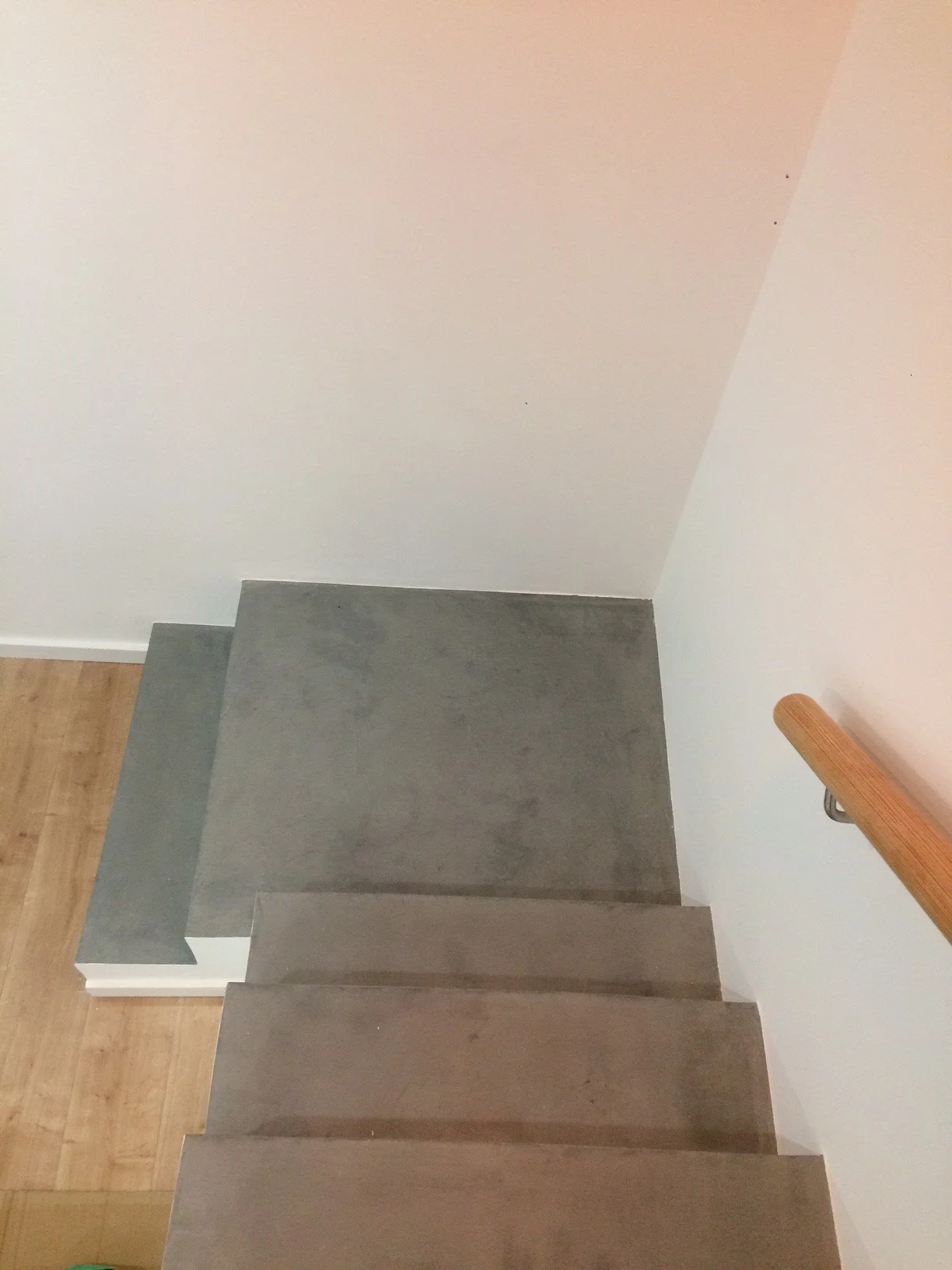 Escada de madeira com degraus de cimento, vista de cima, ao lado de uma parede branca e corrimão de madeira.