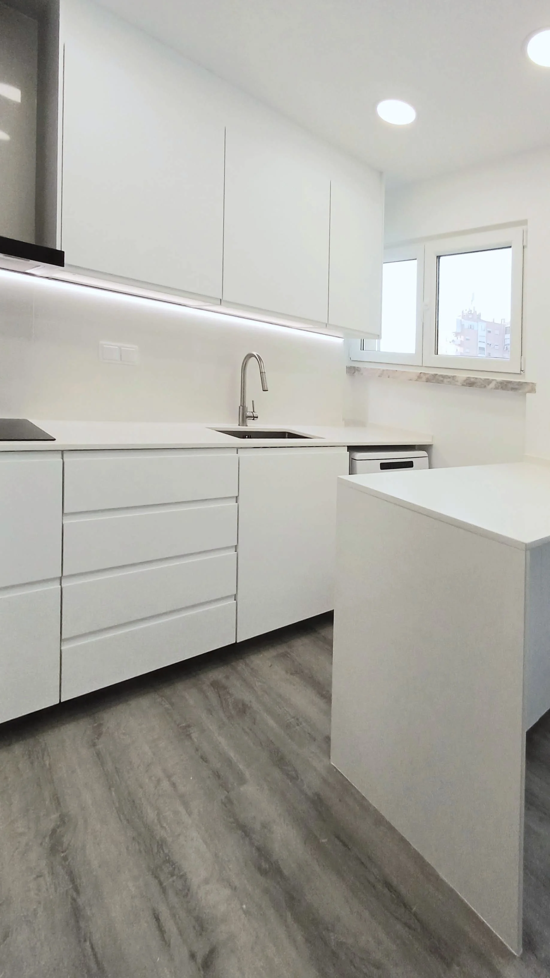 Remodelação de Cozinha no Parque das Nações, Lisboa. Cozinha com Decoração Preto/Branco, Pavimento vinílico imitação de madeira cinza, armários brancos mate com puxador integrado, tampo e back-splash em silstone branco. Equipamentos em cor preta.