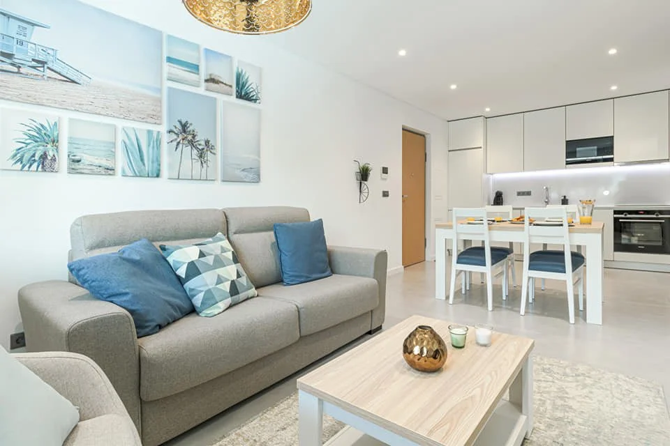 Sala de estar moderna com sofá cinza, almofadas azuis e quadros de praia na parede, incluindo fotos de palmeiras, praia e oceano, com uma cozinha branca ao fundo e uma mesa de jantar, decoração clean e iluminação suave.