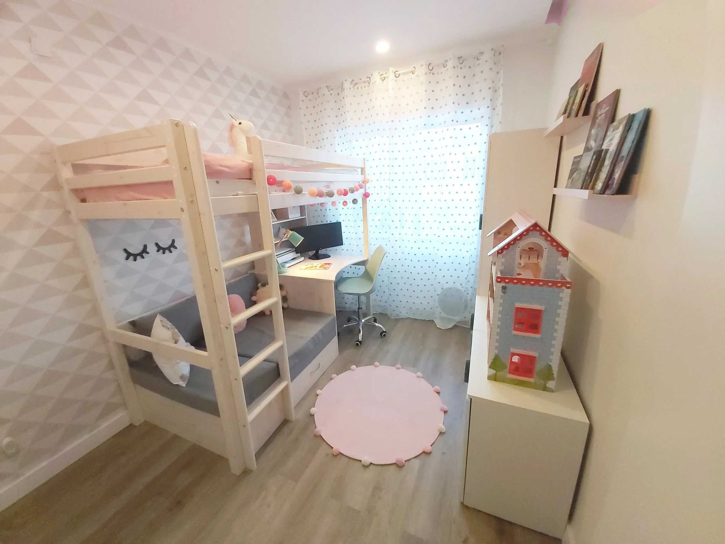 Quarto de criança com cama tipo beliche branca e decorada com uma cabeça de unicórnio de pelúcia, parede com padrão de triângulos, cômoda branca com uma casa de brinquedo no topo, parede com prateleiras e livros, cortina de poás brancas e azuis, tape