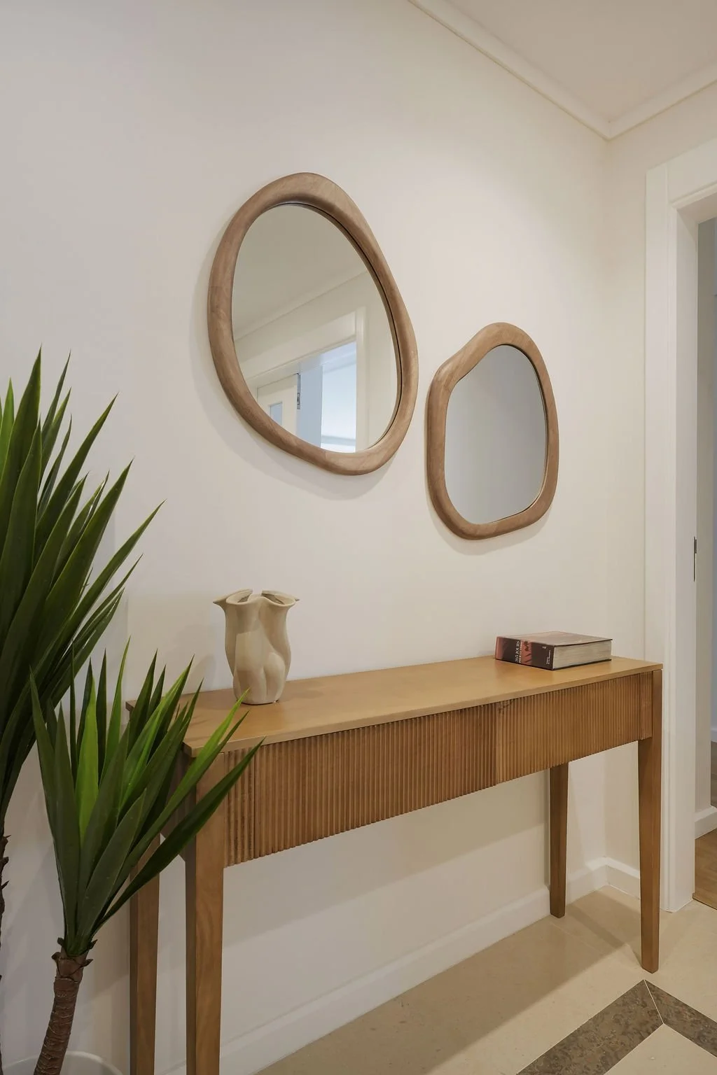 Hall de entrada com móvel de madeira, vaso com planta, dois espelhos de parede de formato irregular de cariz rústico.