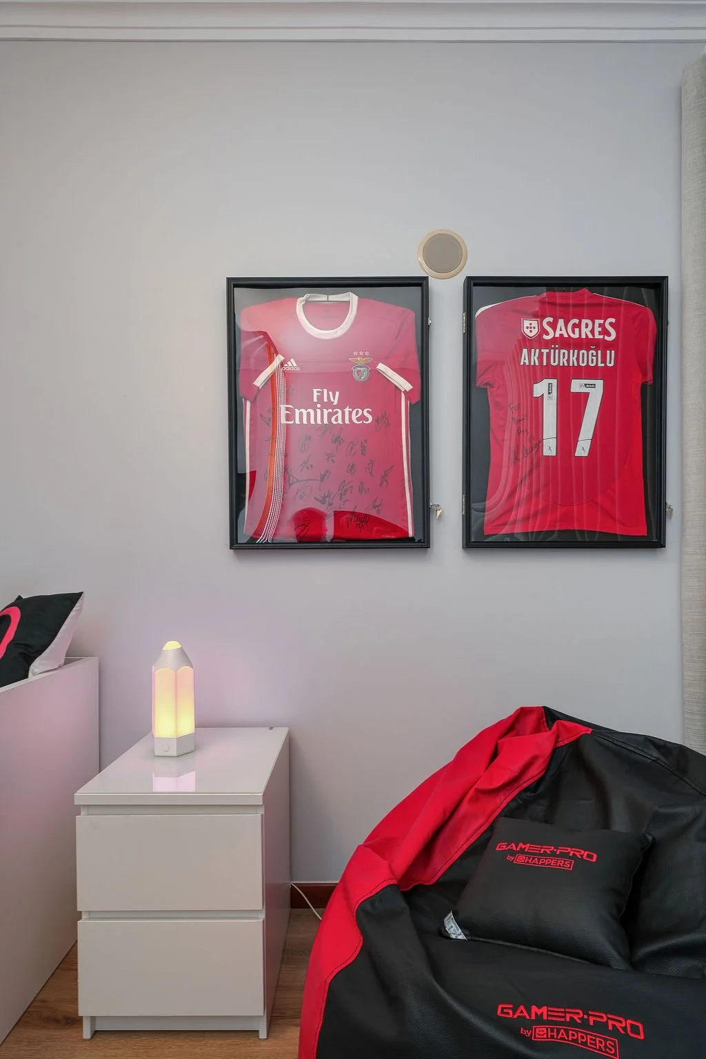 Quartos decorados com camisas de times de futebol FIFA, incluindo uma do Benfica com várias assinaturas e uma do esportista Artur Korgoğlu, as duas estão em quadros na parede. A foto também mostra uma cama, uma luminária e um saco de dormir na cor pr