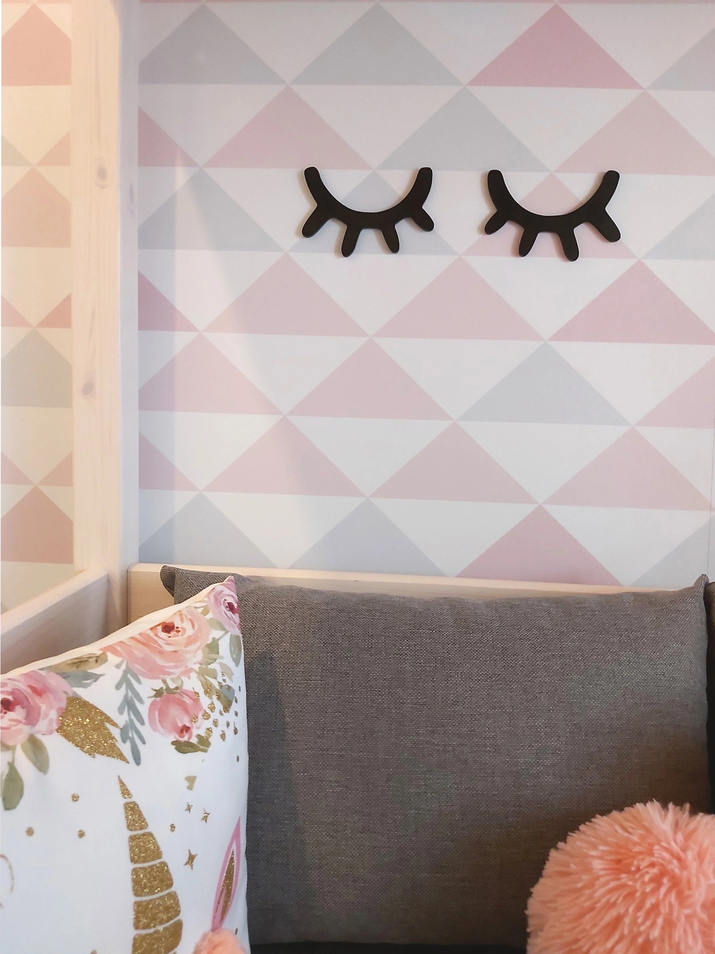 Decoração de uma parede com dois olhos de mentira em formato de 'blush' com cílios grandes e curvos, ao lado de um sofá com almofadas coloridas, incluindo uma com estampa de unicornio e glitter dourado, e uma almofada com pelúcia rosa, criando um amb