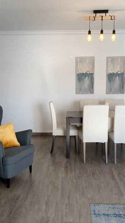 Sala de jantar com mesa retangular branca, seis cadeiras brancas ao redor, sofá cinza com travesseiro amarelo ao lado, luz pendurada com três lâmpadas, paredes com duas obras de arte abstratas.