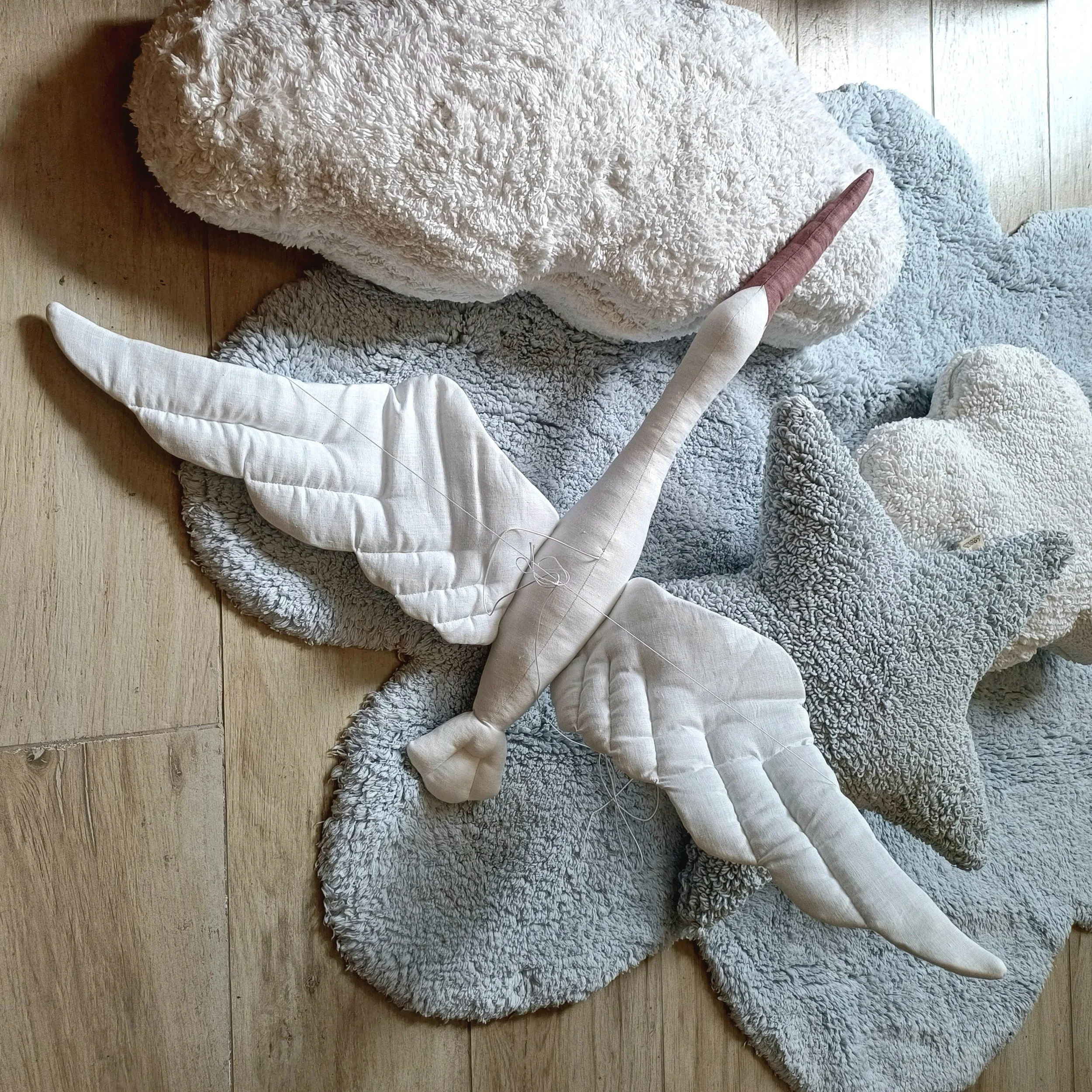 Decoração de festa com tema de unicórnio, incluindo uma asa de pano branca, um chifre rosa e cabeça de unicórnio em tecido, e nuvens de pelúcia cinza e branca, tudo colocado sobre um tapete cinza em um piso de madeira.