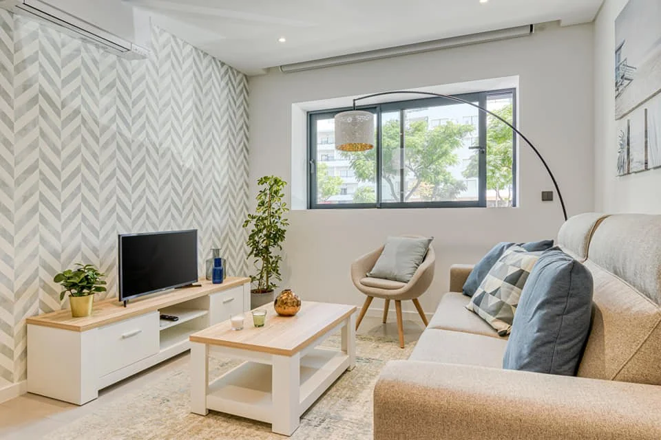 Home Staging de Apartamento em Quarteira, Algarve.
Sala de estar moderna com sofá cinza, almofadas azuis e quadros de praia na parede, incluindo fotos de palmeiras, praia e oceano. Aplicação de papel em parede de TV. Decoração clean num ambiente repl
