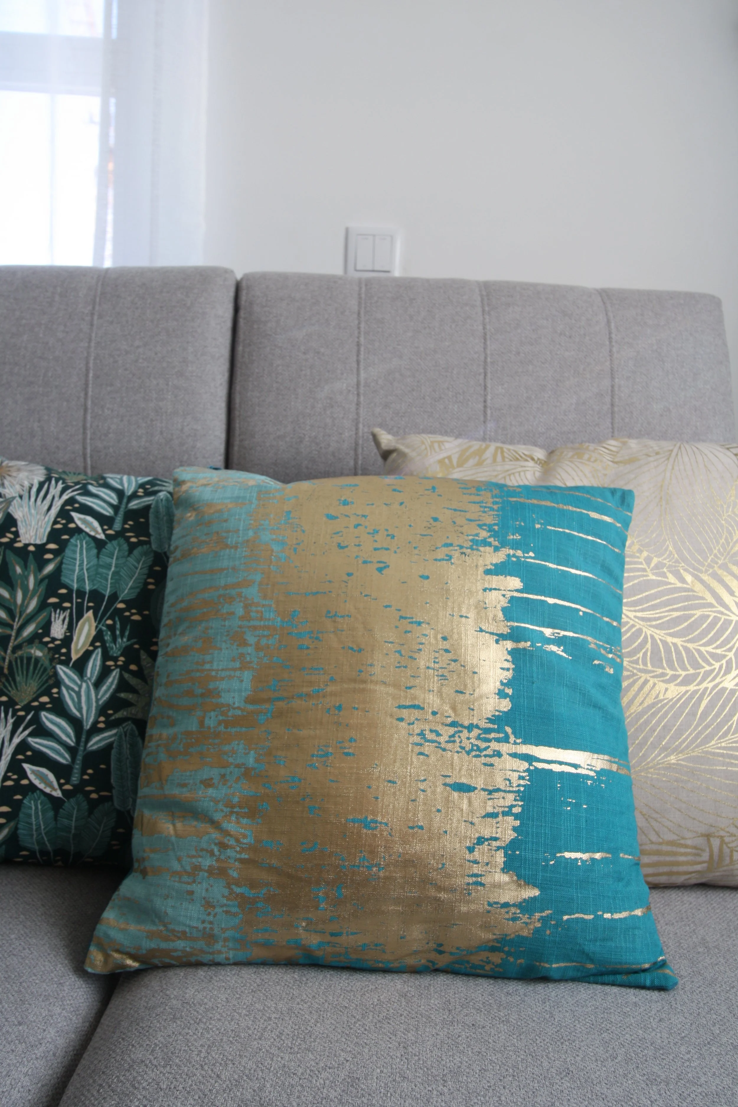 Três almofadas decorativas em sofá cinza claro, uma com padrão de folhas verdes escuras, uma azul com detalhes dourados, e outra com estampa de folhas douradas.