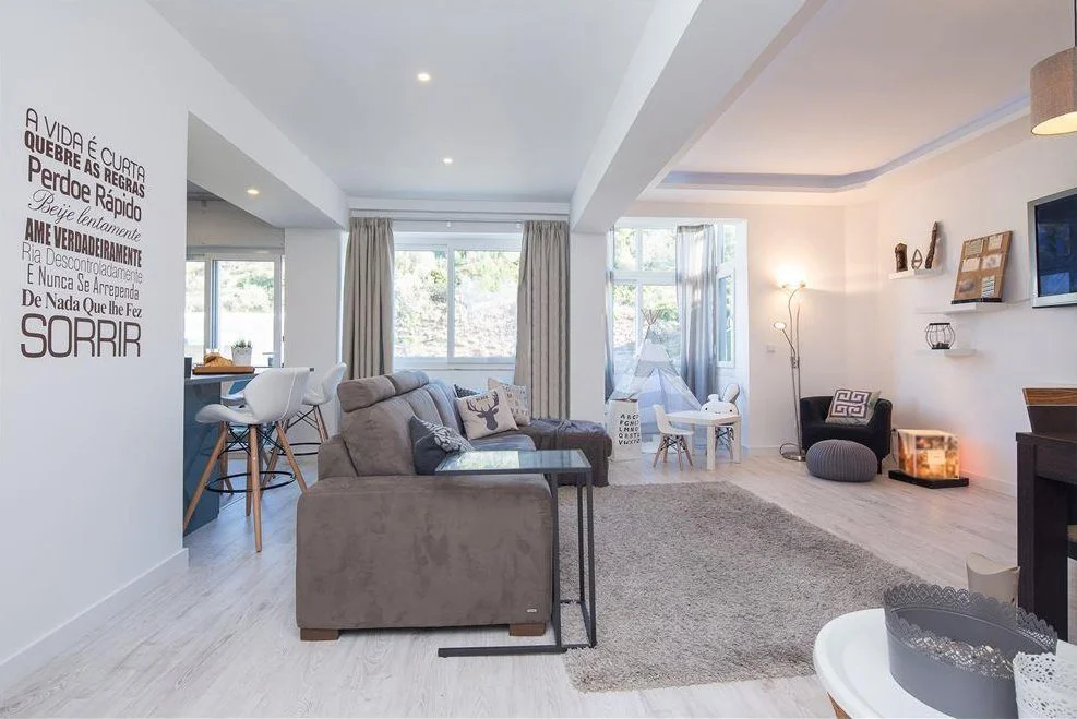 Sala de estar com sofá cinza, tapete bege, cadeira preta, televisores na parede, janelas grandes com cortinas, ambiente bem iluminado e decorado com objetos de decoração