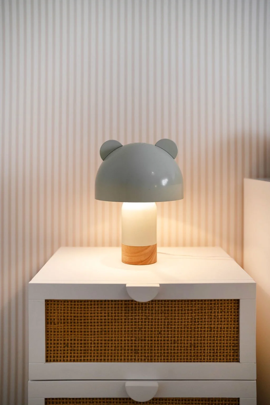 Luminária com gorro de ursinho em cima de uma cômoda branca com tampo de madeira, parede com papel de parede listrado ao fundo.
