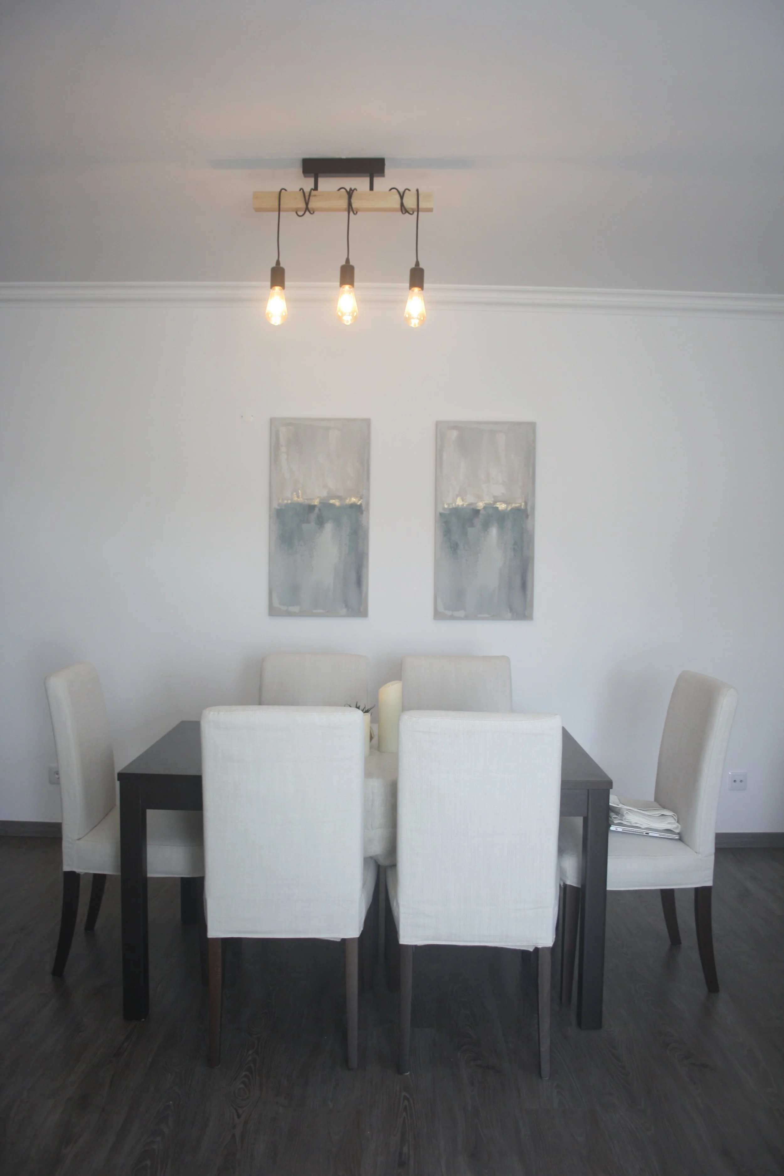 Sala de jantar com mesa preta, seis cadeiras brancas, duas obras de arte abstratas na parede, luminária com três lâmpadas de filamento no teto.