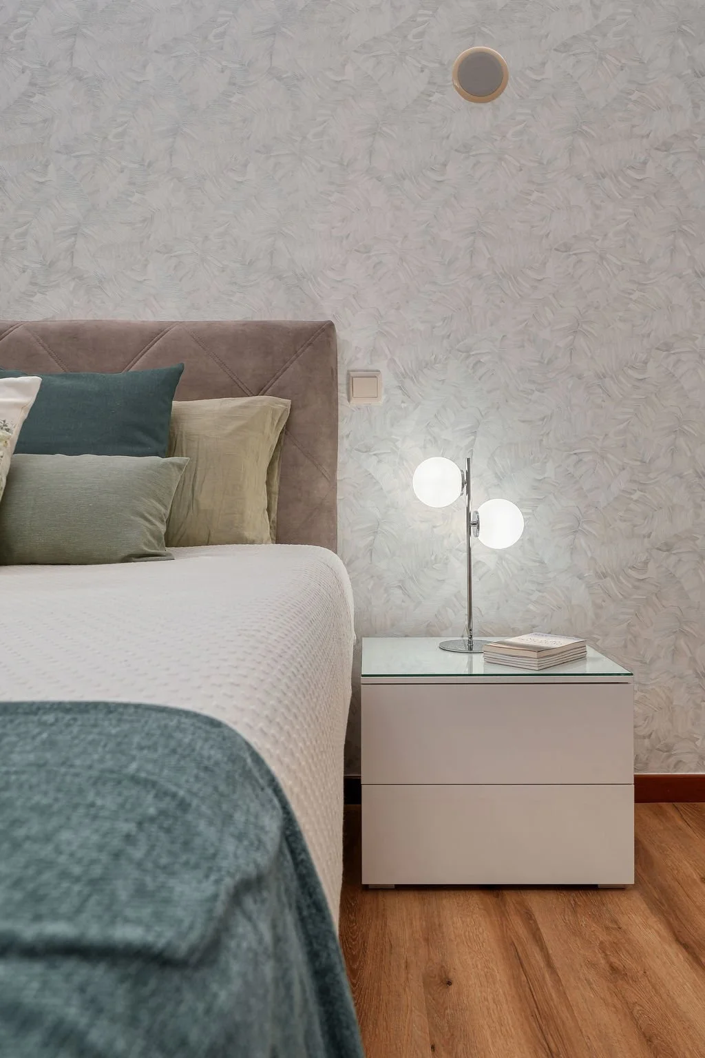 Quarto com cabeceira estofada rosa claro, cama com colchas brancas e almofadas em tons neutros e azul. Ao lado, uma mesa de cabeceira branca com duas gavetas, luminária com dois globos brancos e um livro.