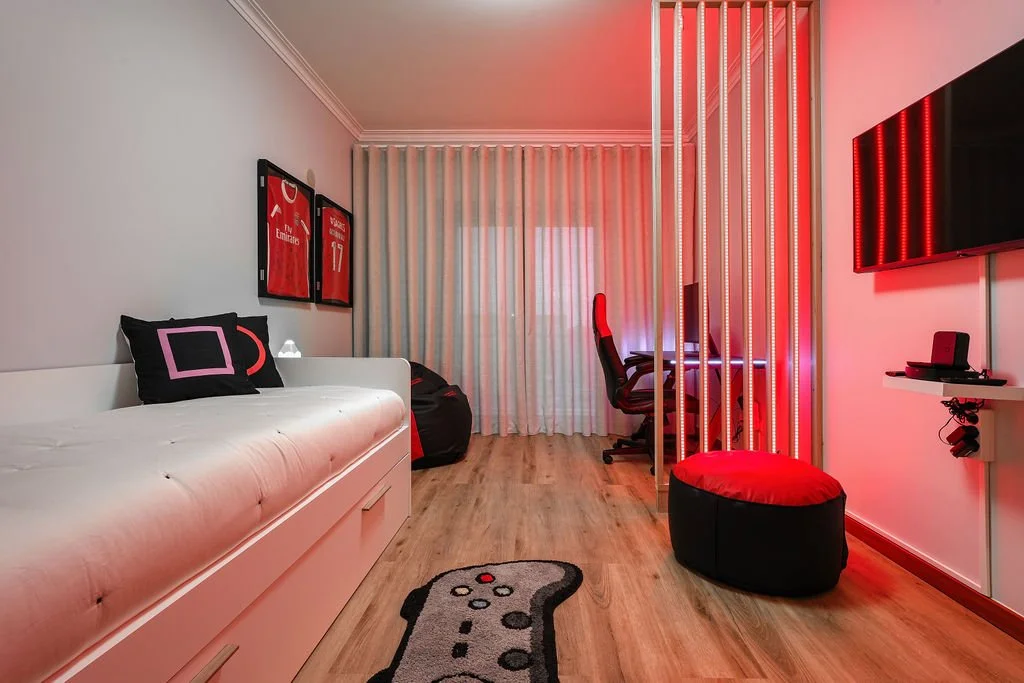 Quarto organizado com paredes brancas, cortinas brancas, sofá branco com travesseiros pretos com detalhes em rosa, quadro de camisa de futebol pendurado na parede, piso de madeira, tapete com controle de videogame, cadeira de escritório, pufe vermelh