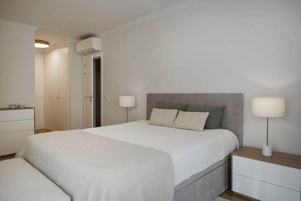 Quarto com cama de casal, cabeceira cinza, duas luminárias de mesa, guarda-roupa embutido, ar-condicionado e decoração minimalista em tons neutros.