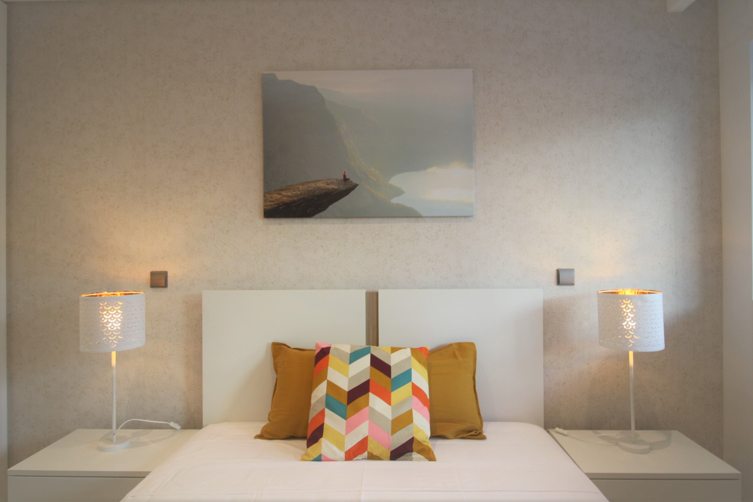 Quarto com cama de casal, almofadas coloridas e de cor marrom, com duas luminárias de mesa ao lado da cama, e uma pintura de paisagem na parede acima da cama