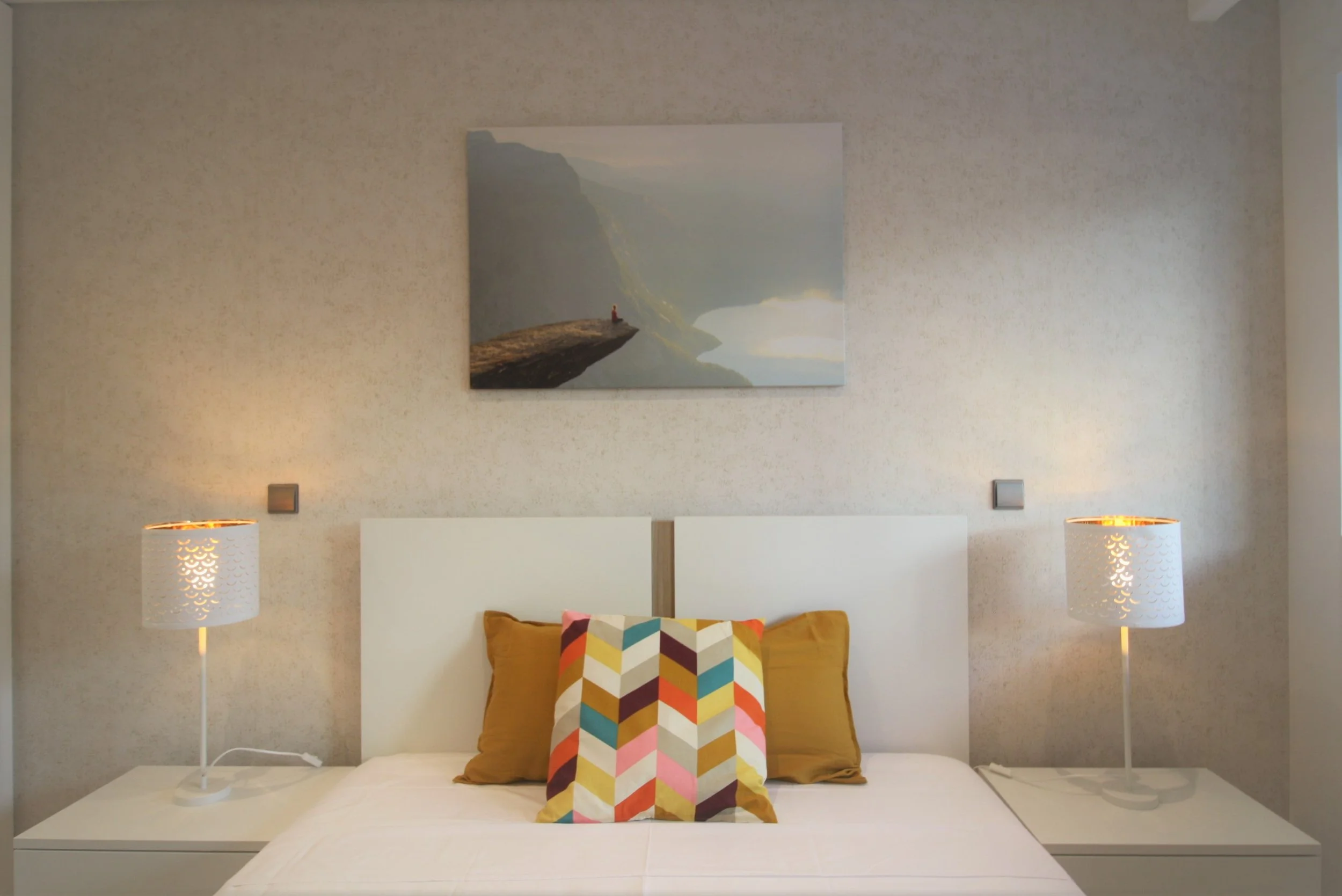 Home Staging de Apartamento em Quarteira, Algarve.
Quarto com casal decorado com almofadas coloridas e de cor mostarda, com dois candeeiros de mesa. Quadro  de paisagem suspenso em parede revestida a papel de parede.