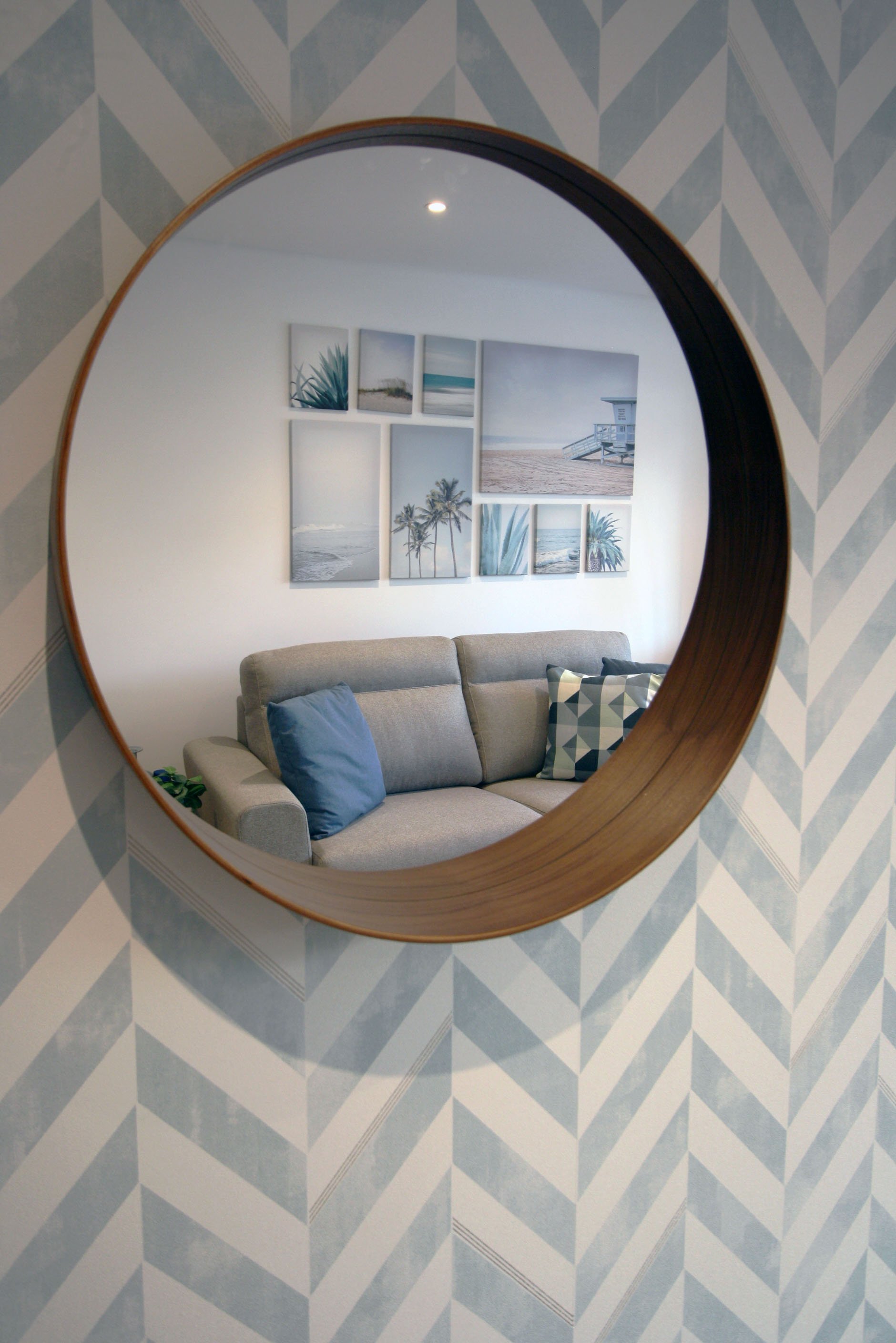 Espelho redondo refletindo uma sala com sofá cinza, almofadas azul e com padrão de triângulos, parede branca com quadros de fotos de praia, e parede com papel de parede com padrão geométrico azul e branco.