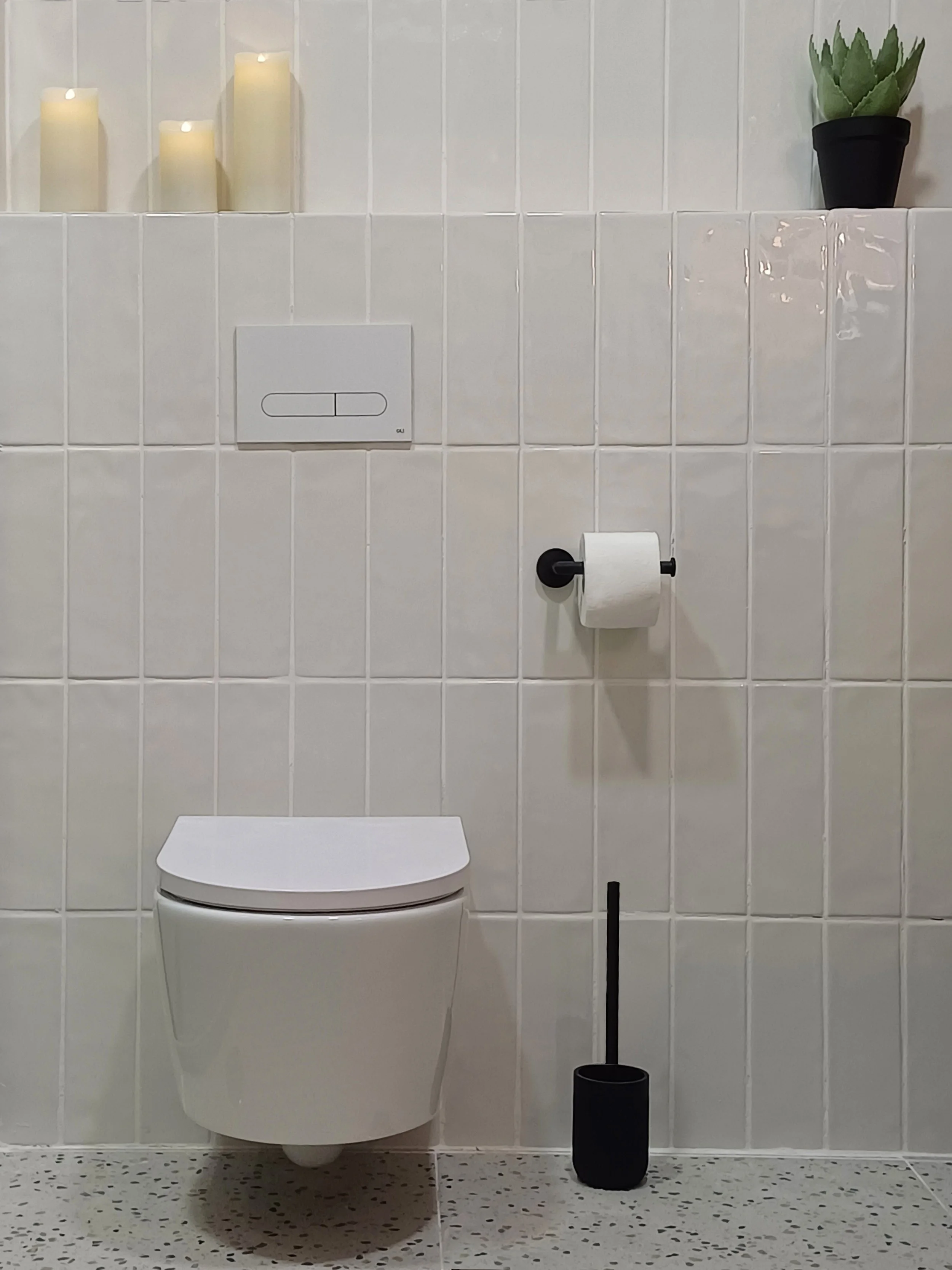 Banheiro com vaso sanitário branco, papel higiênico na parede e uma escova sanitária preta ao lado. Decorado com velas e uma planta suculenta na prateleira