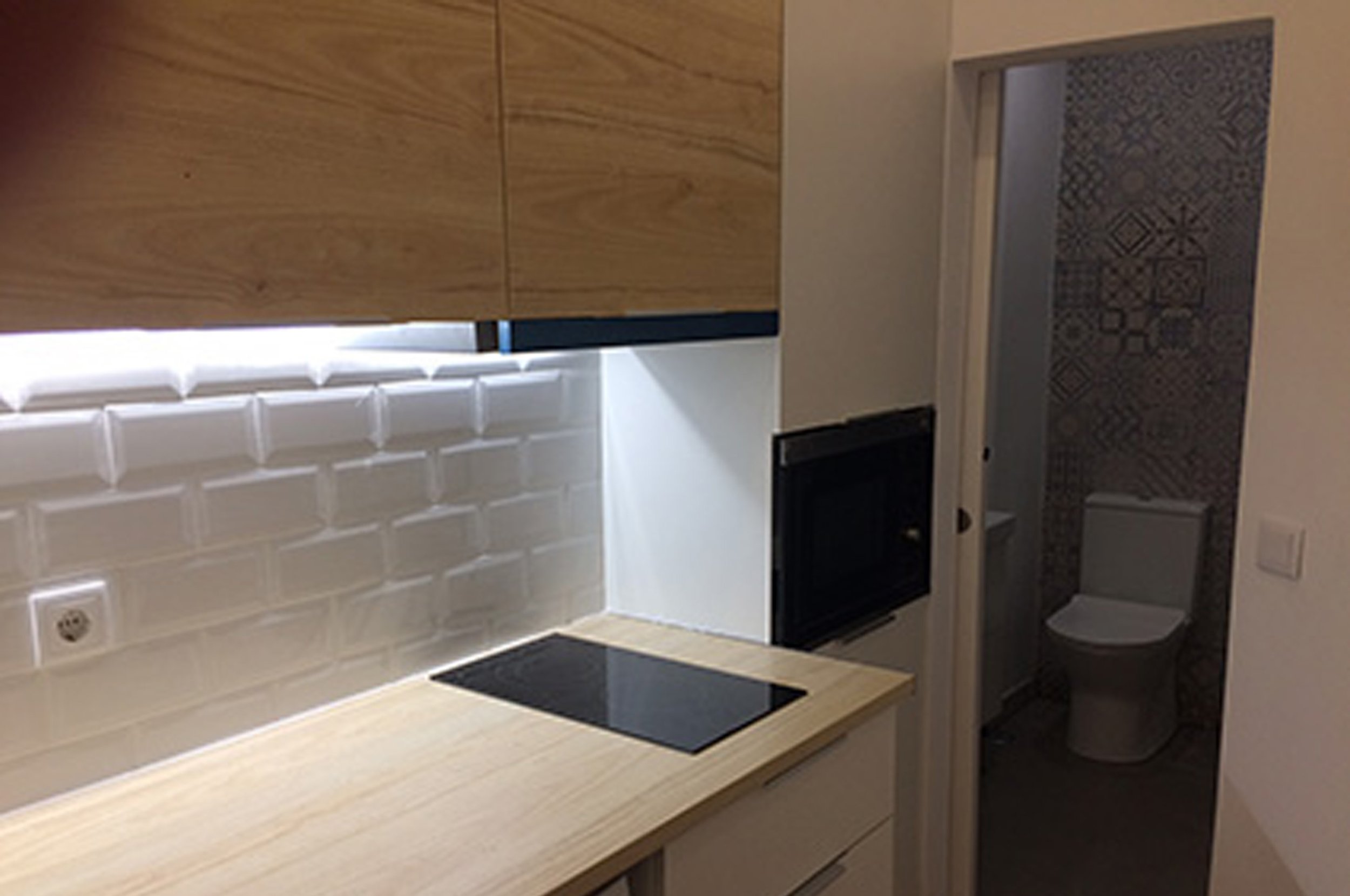 Cozinha com bancada de madeira clara, backsplash de azulejos brancos, forno embutido e uma porta que leva a um banheiro com vaso sanitário.